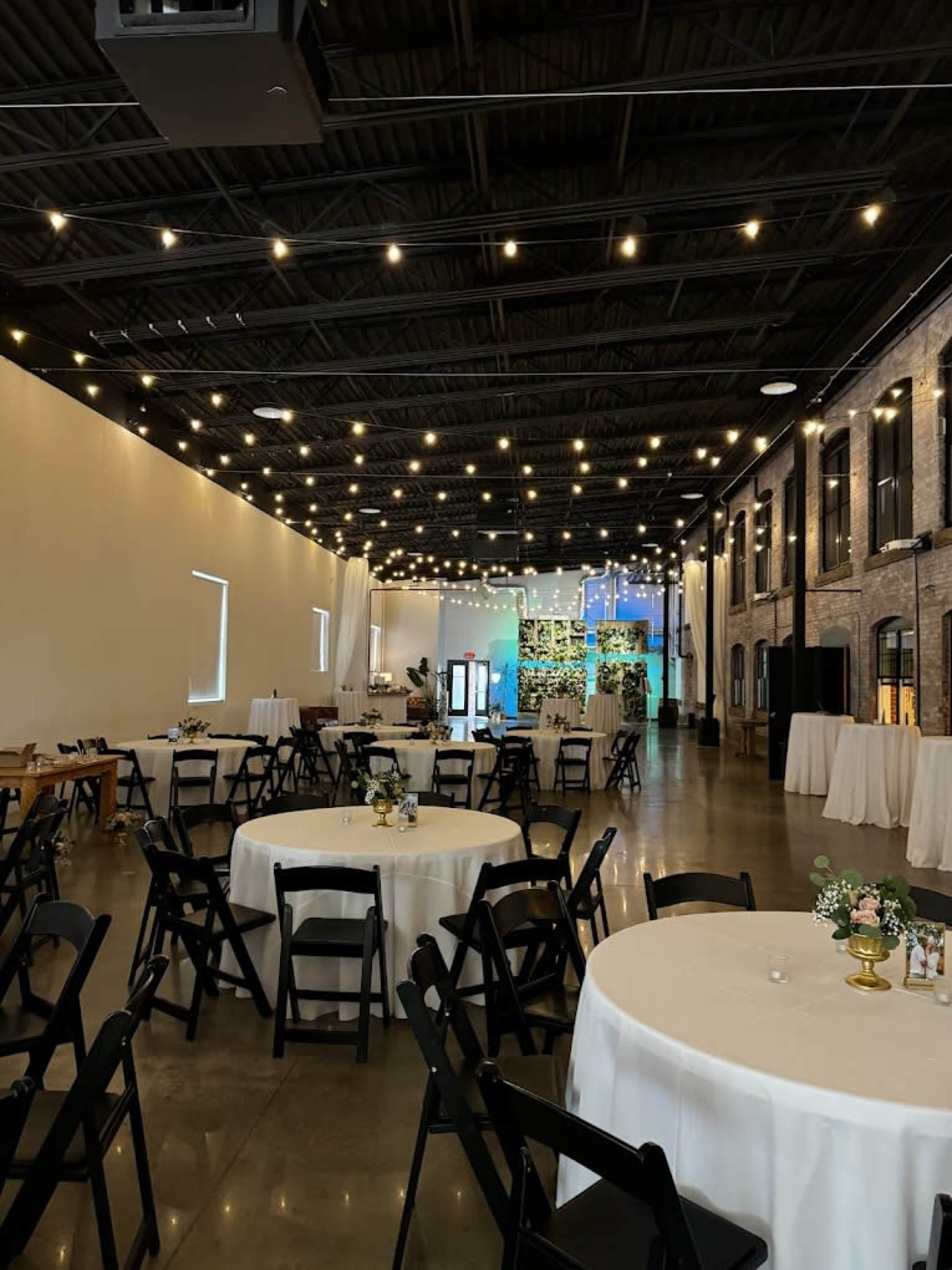 The image shows a spacious event venue with round tables set for dining, string lights overhead, and an elegantly decorated stage at the far end.