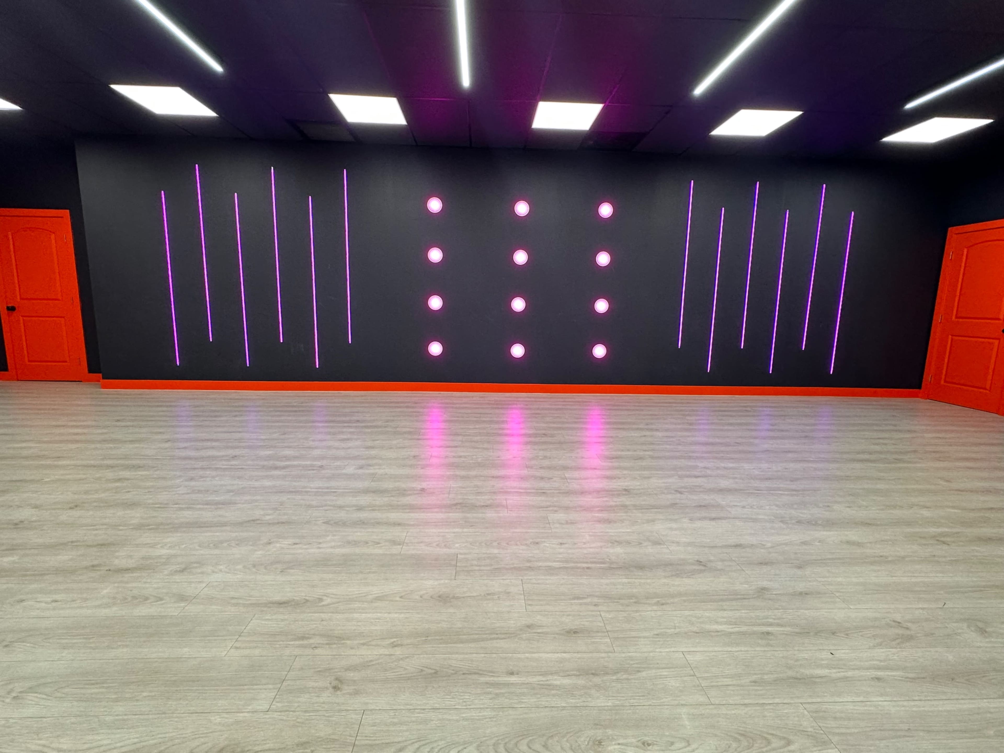 A spacious interior room with a dark wall featuring vertical neon lights, orange doors, and a light wooden floor.