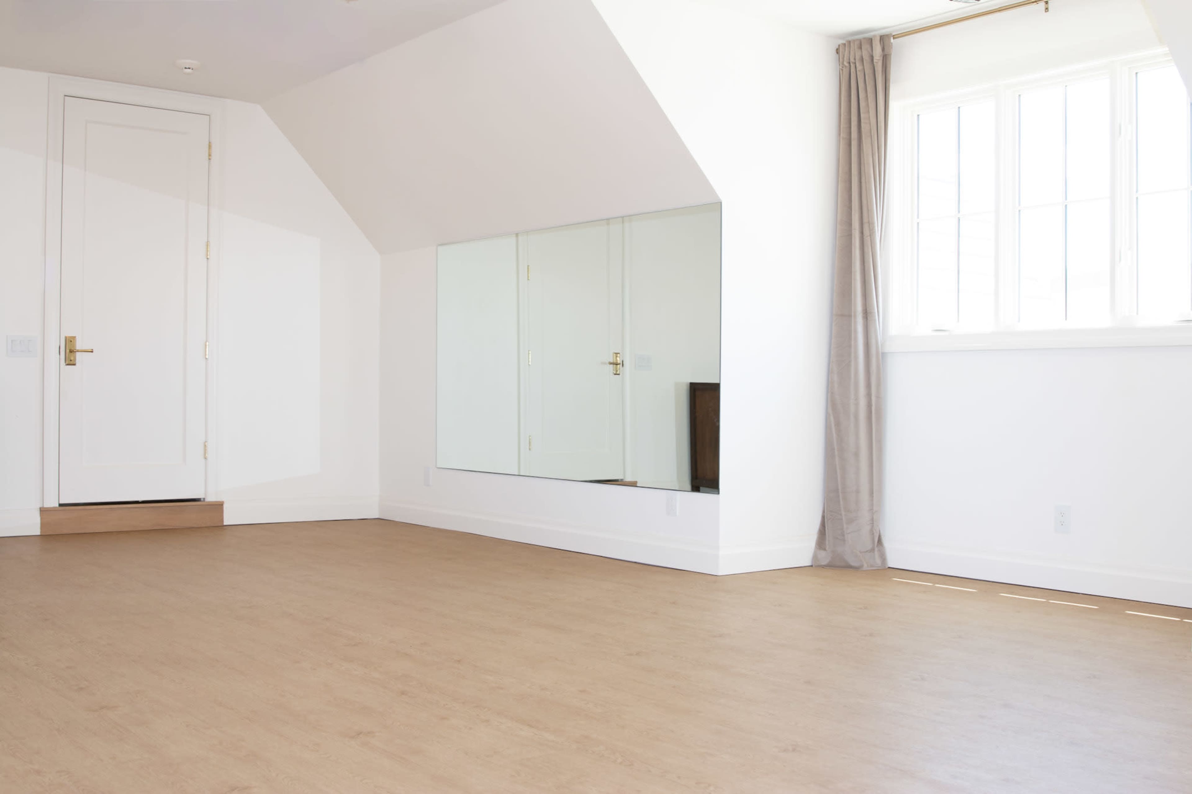 The room features a large, empty space with a wooden floor, a door, and a mirrored wall opposite a window with natural light.