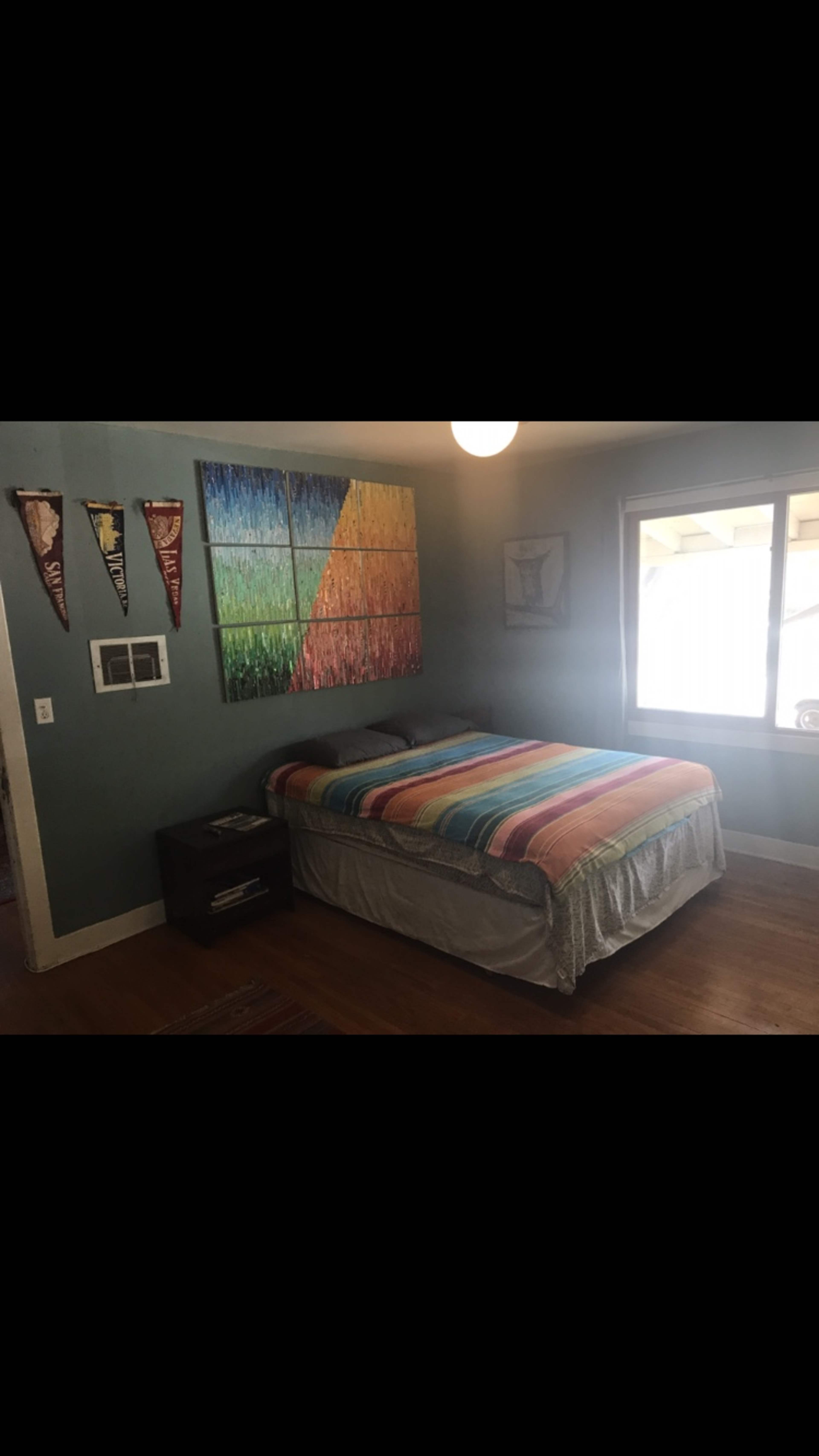 A bedroom features a bed with a multicolored striped blanket, a large abstract painting on the wall, and a window allowing natural light to enter.
