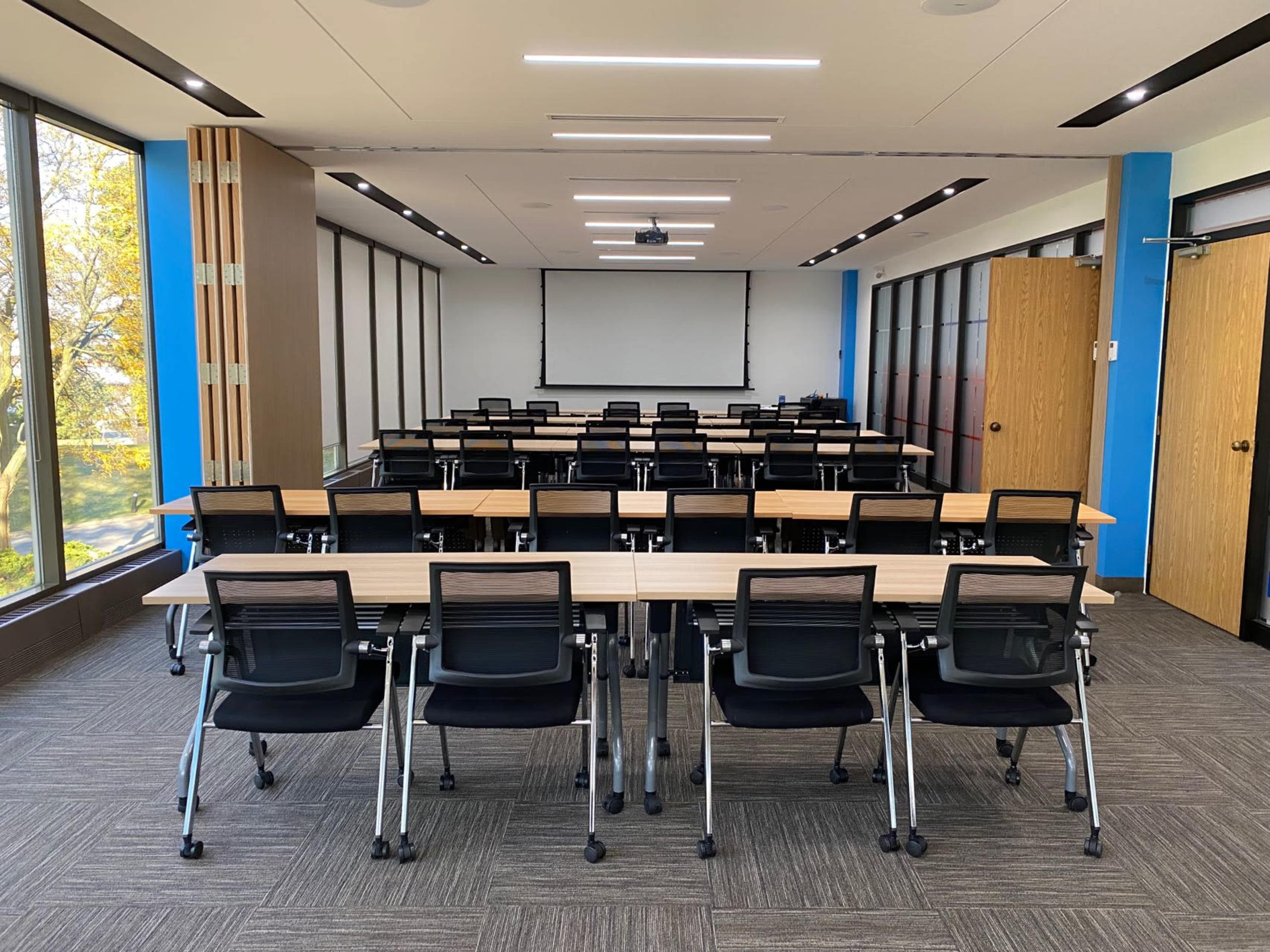 A modern meeting room features rows of tables and black chairs with large windows providing natural light.