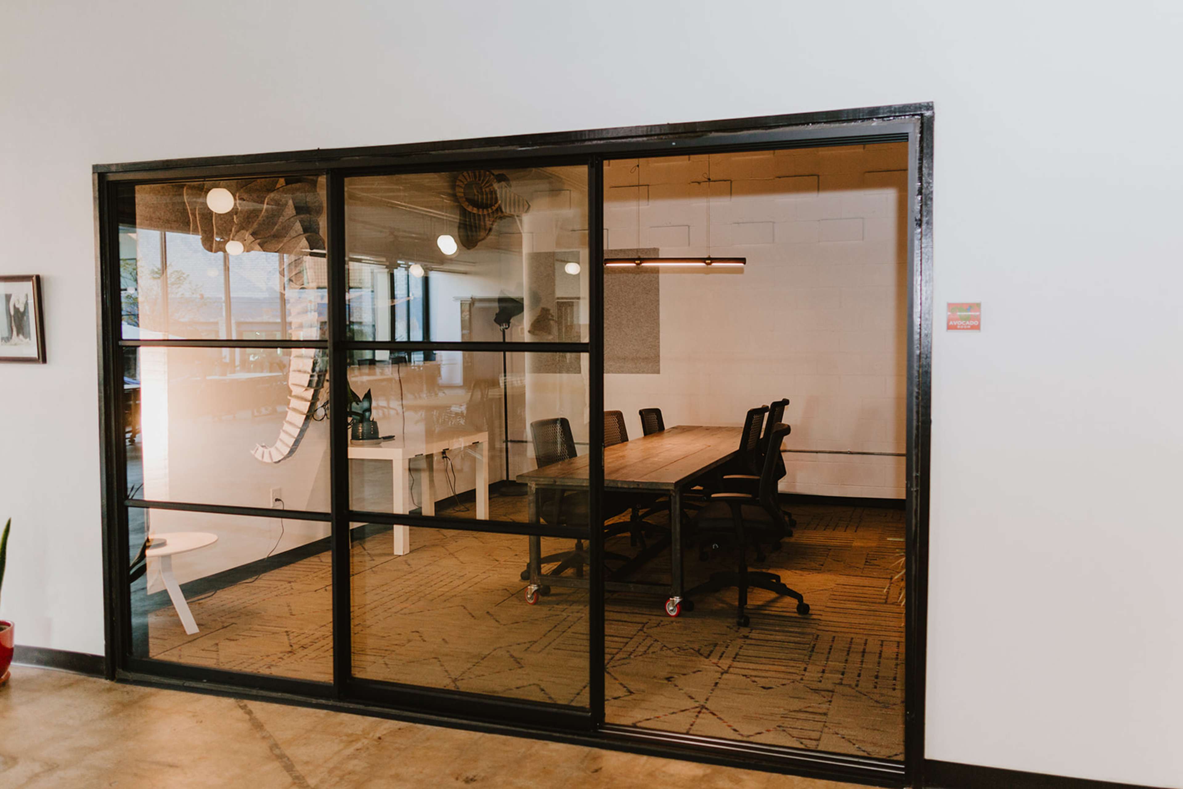 A modern conference room features a large wooden table surrounded by black chairs, enclosed by a glass wall.
