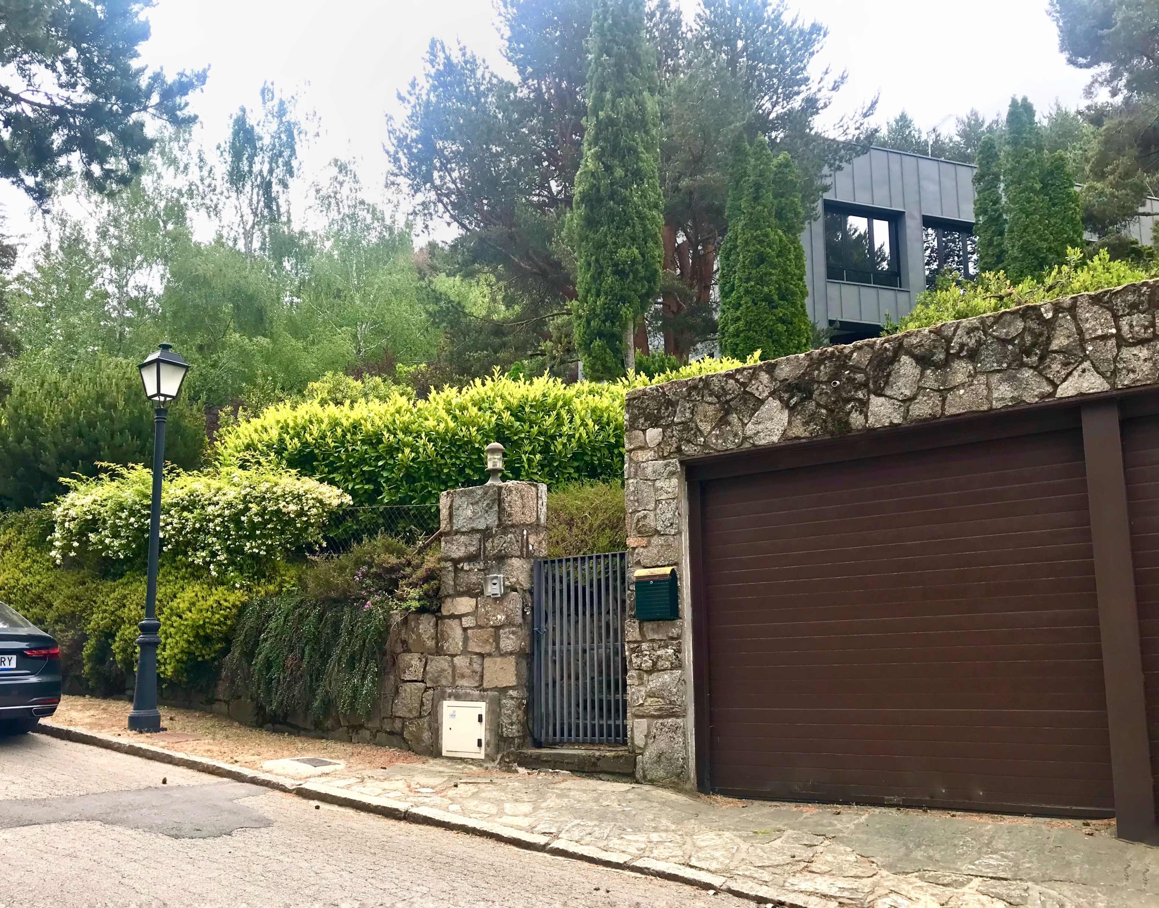 A modern house is set behind a stone wall and gate, surrounded by tall greenery and trees.
