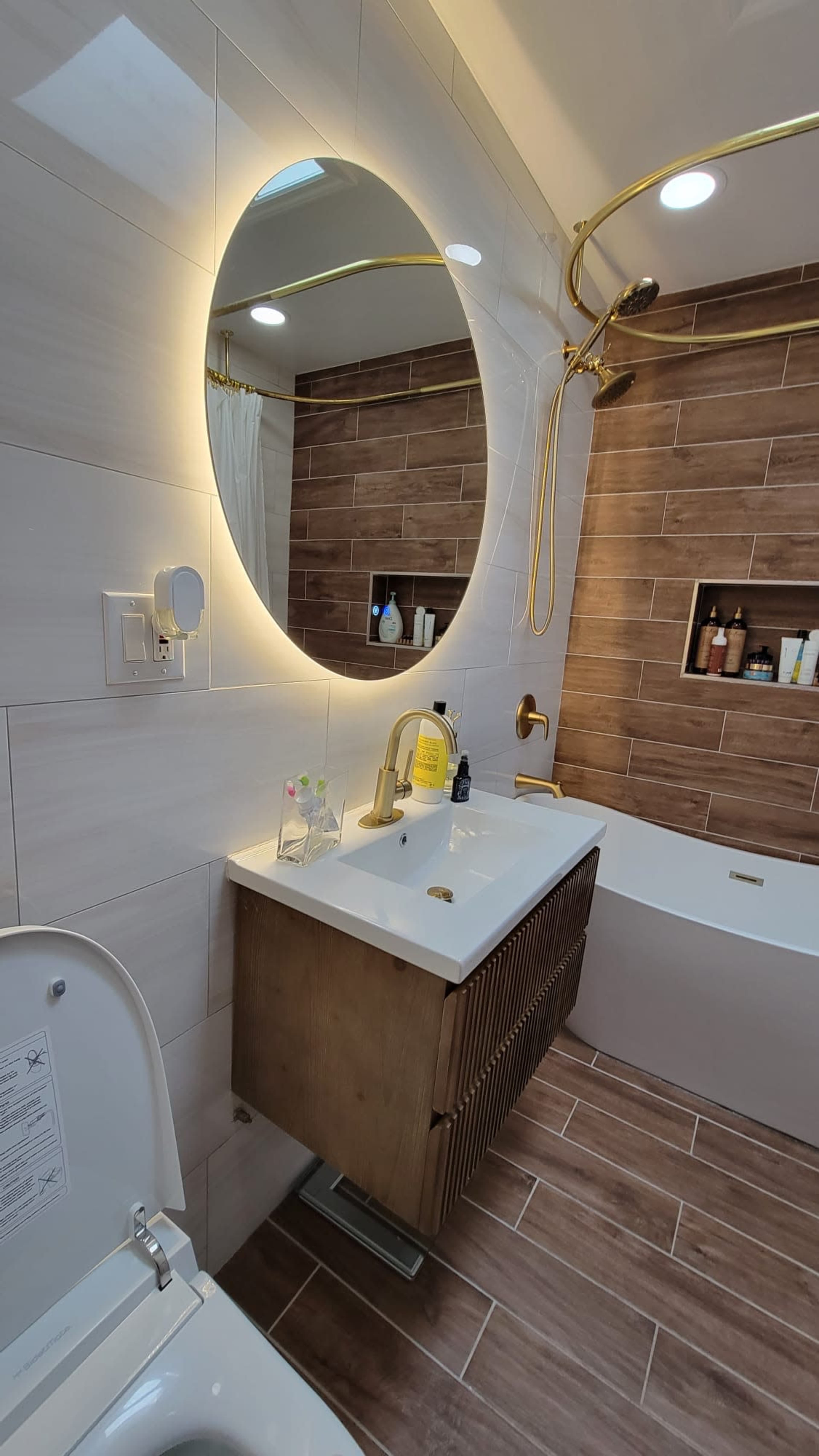 A modern bathroom features a round mirror with backlighting, a gold faucet, and wooden accents on the vanity, with a bathtub and a towel rack in the background.