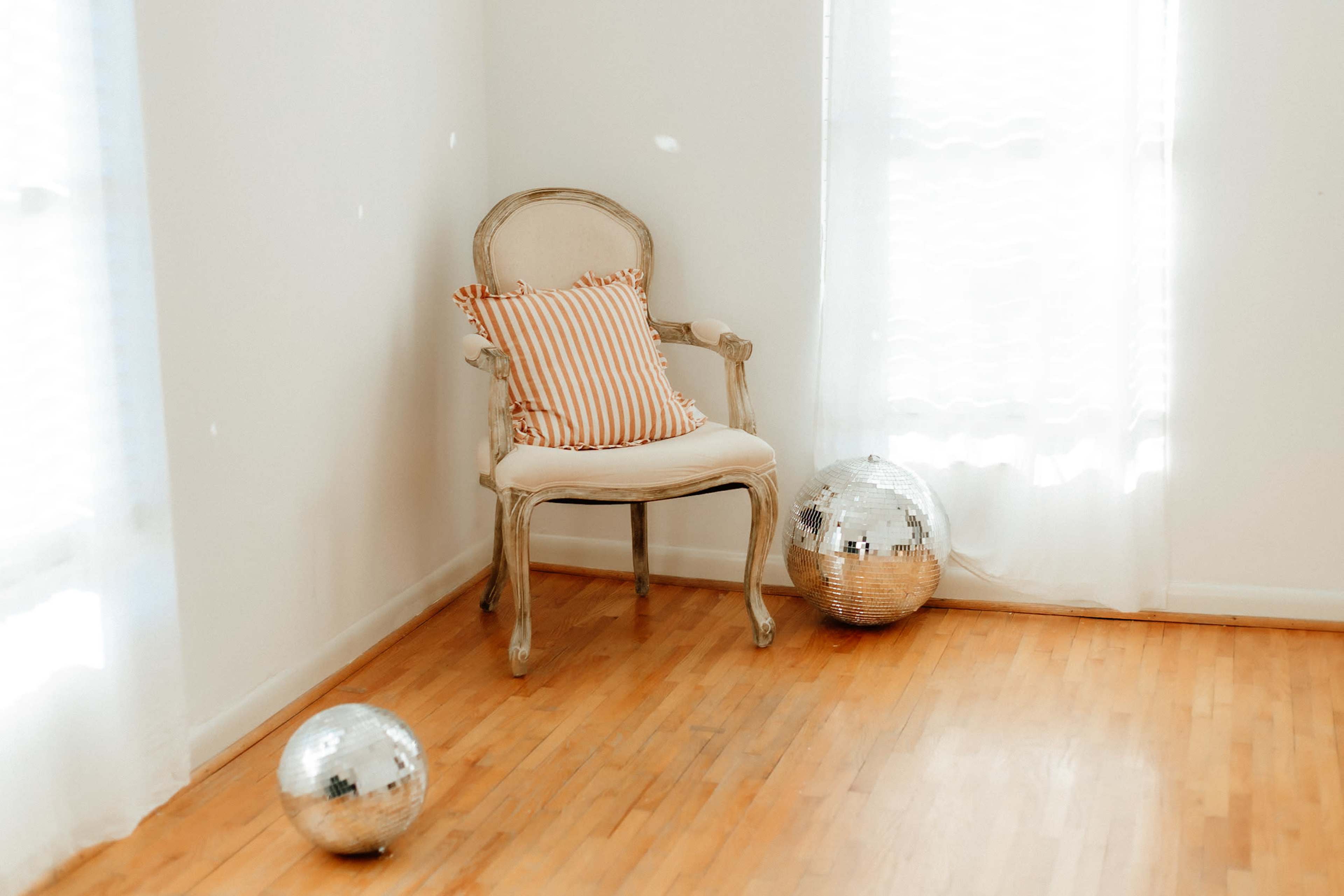 A white-walled room features a single ornate chair with a striped cushion and two disco balls on the wooden floor.