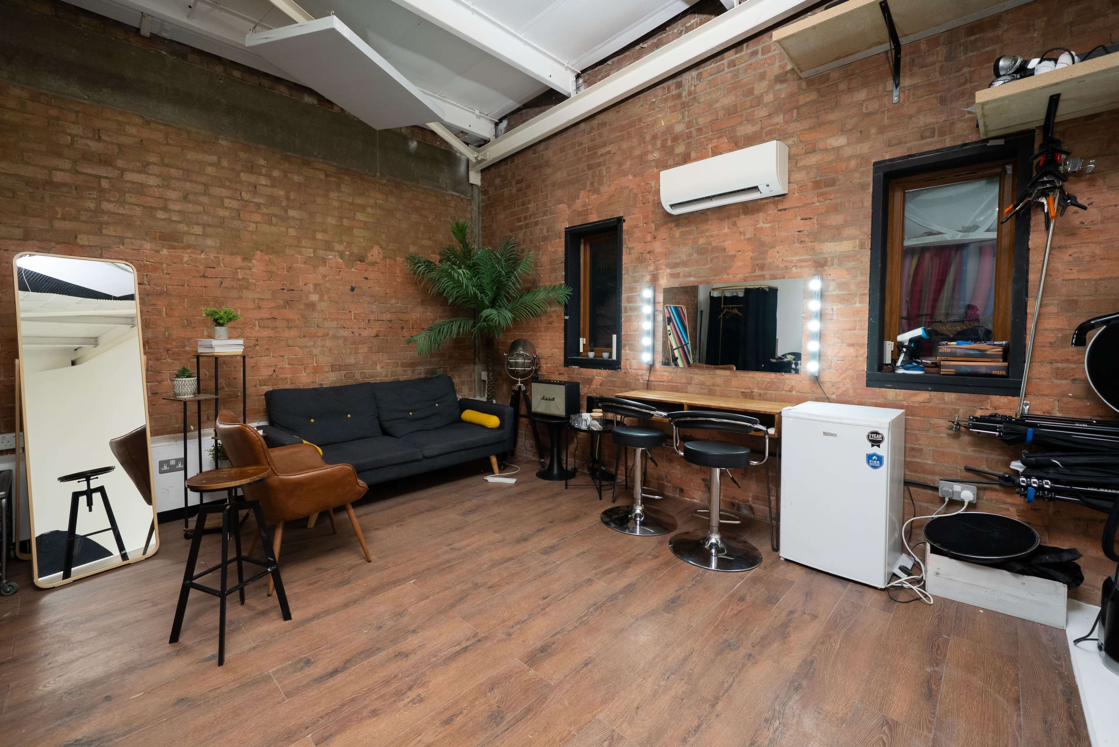 The image shows a modern interior space with exposed brick walls, a sofa, two bar stools at a counter, a full-length mirror, and various furnishings, including a small refrigerator and decorative plants.