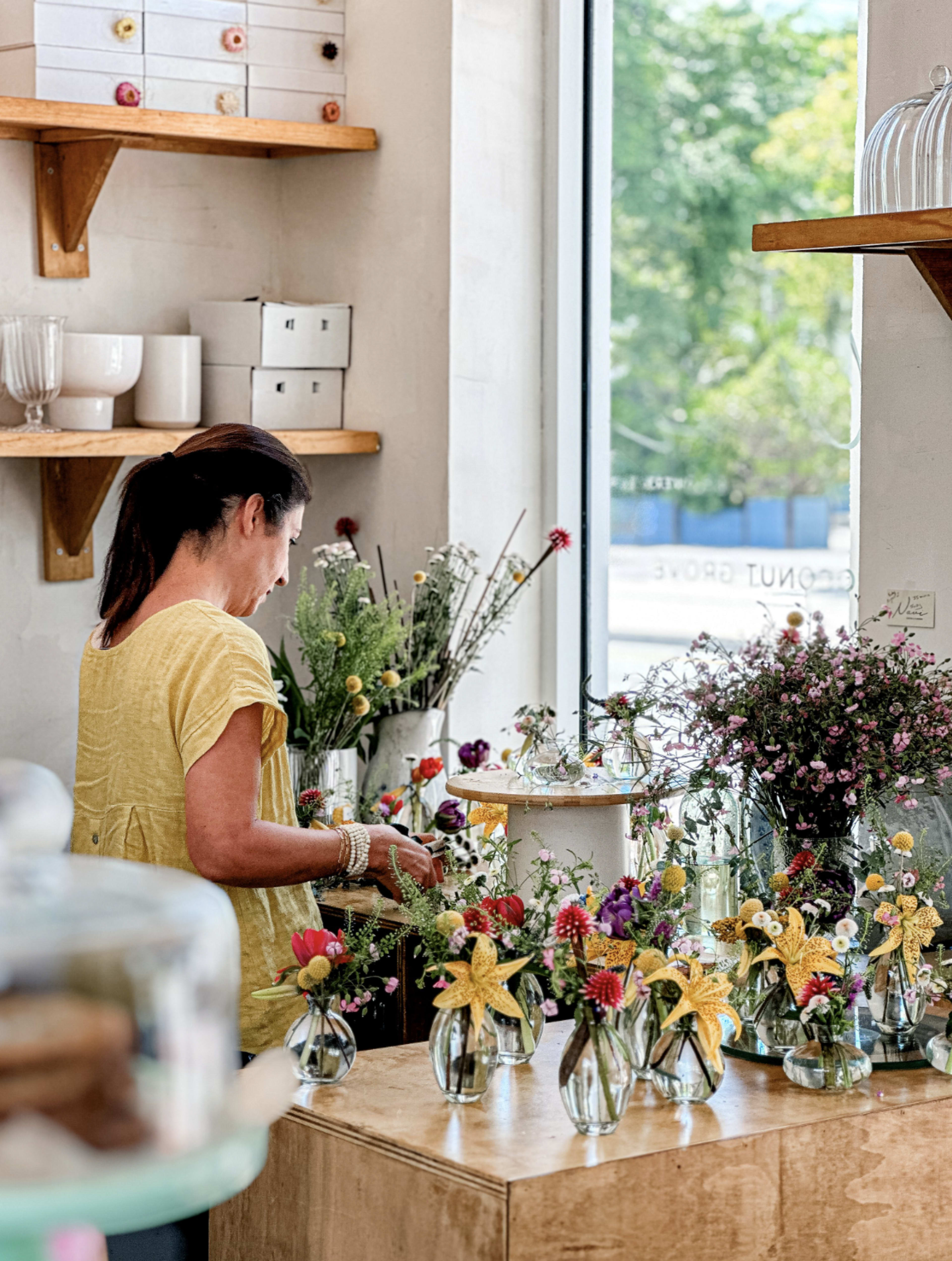A woman in a yellow shirt arranges colorful flowers in small glass vases within a flower shop.