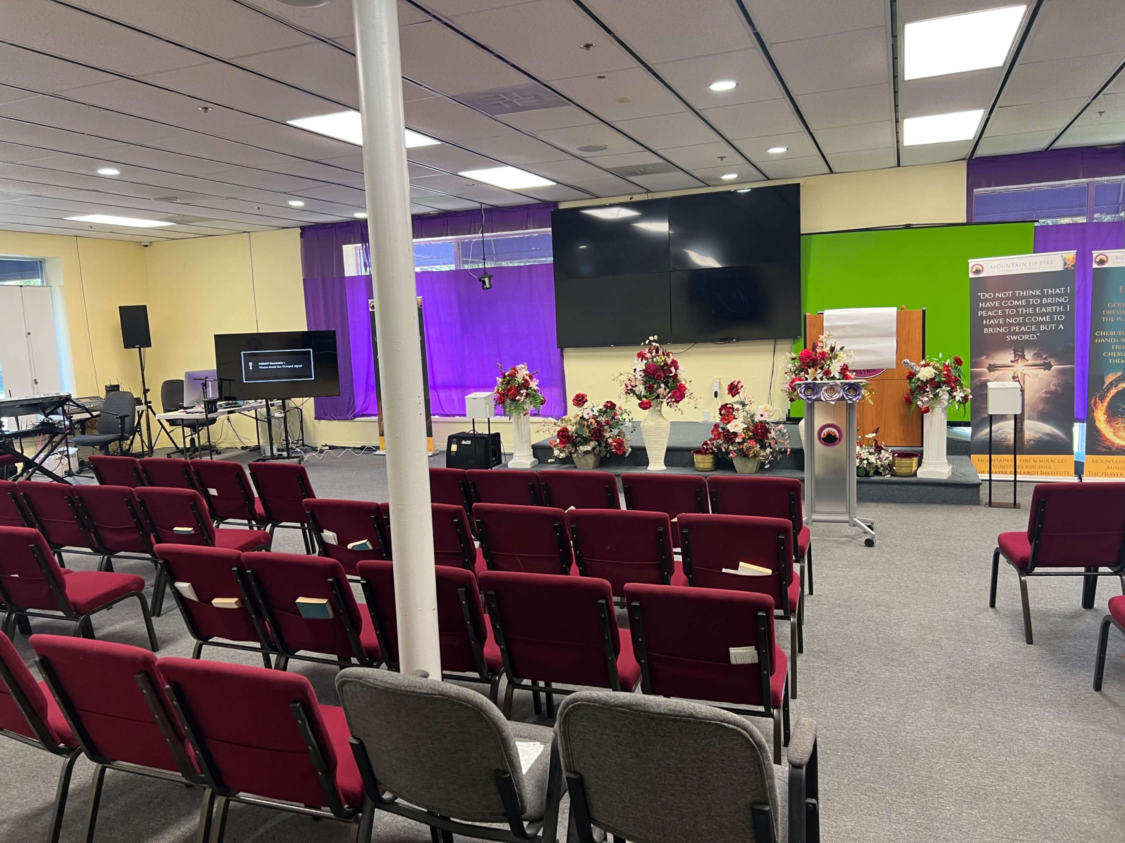 The image shows a brightly lit indoor space arranged with red and gray chairs, a podium with floral decorations, a large screen, and promotional banners on the walls.