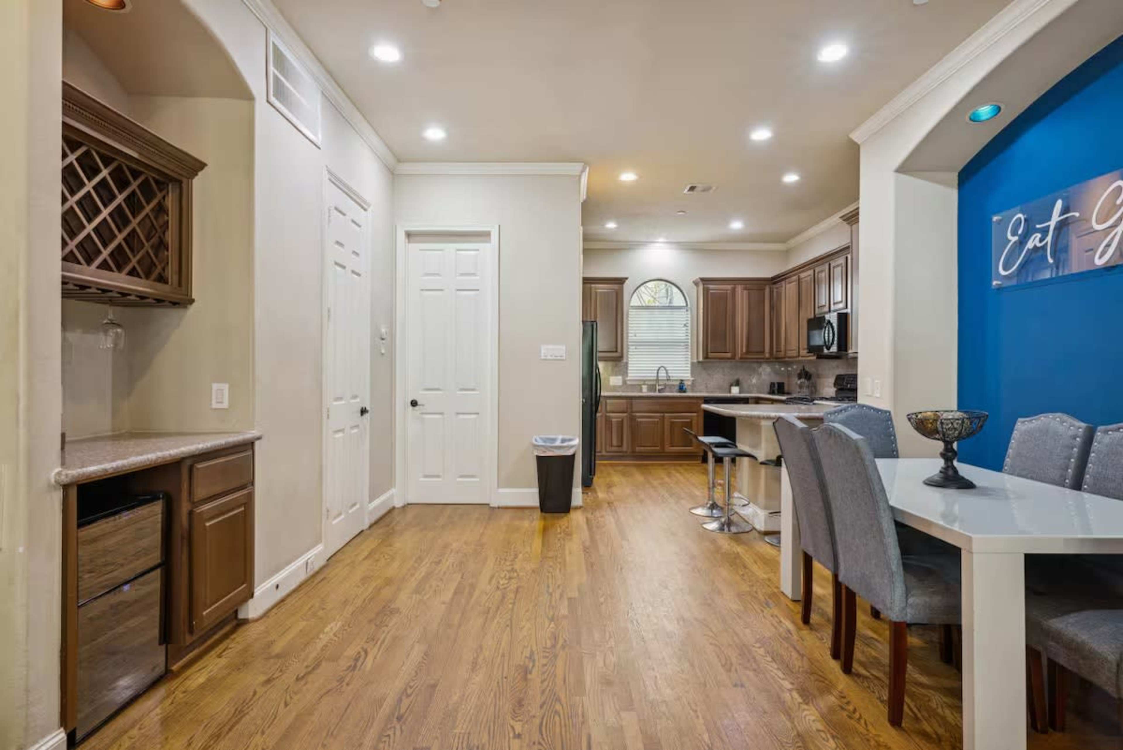 The image shows a modern kitchen and dining area with hardwood flooring, featuring dark cabinetry, a dining table with gray chairs, and a framed sign on the wall.