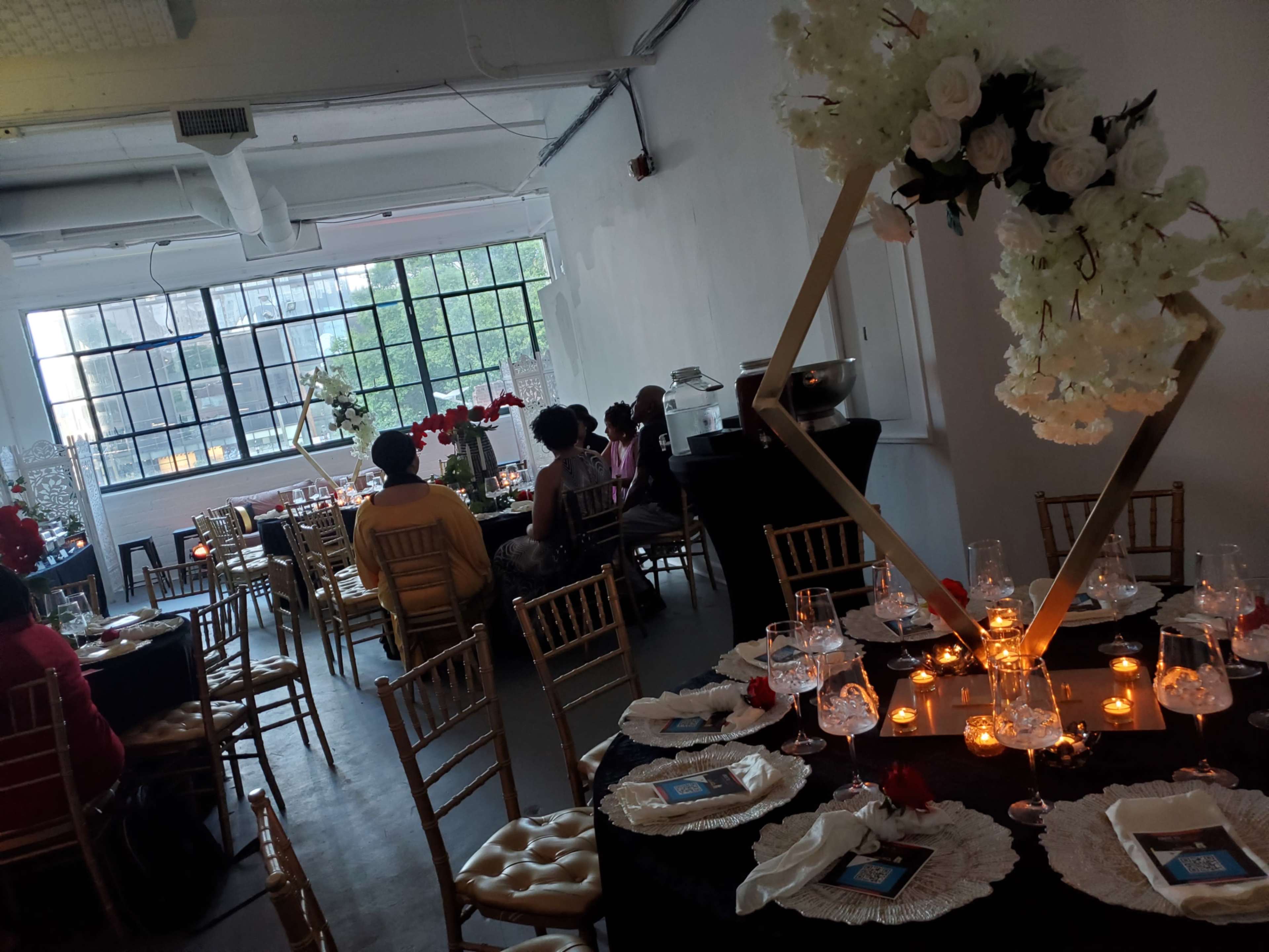 The image shows a decorated event space with tables set for dining, featuring floral arrangements and candles, while guests are seated and socializing.