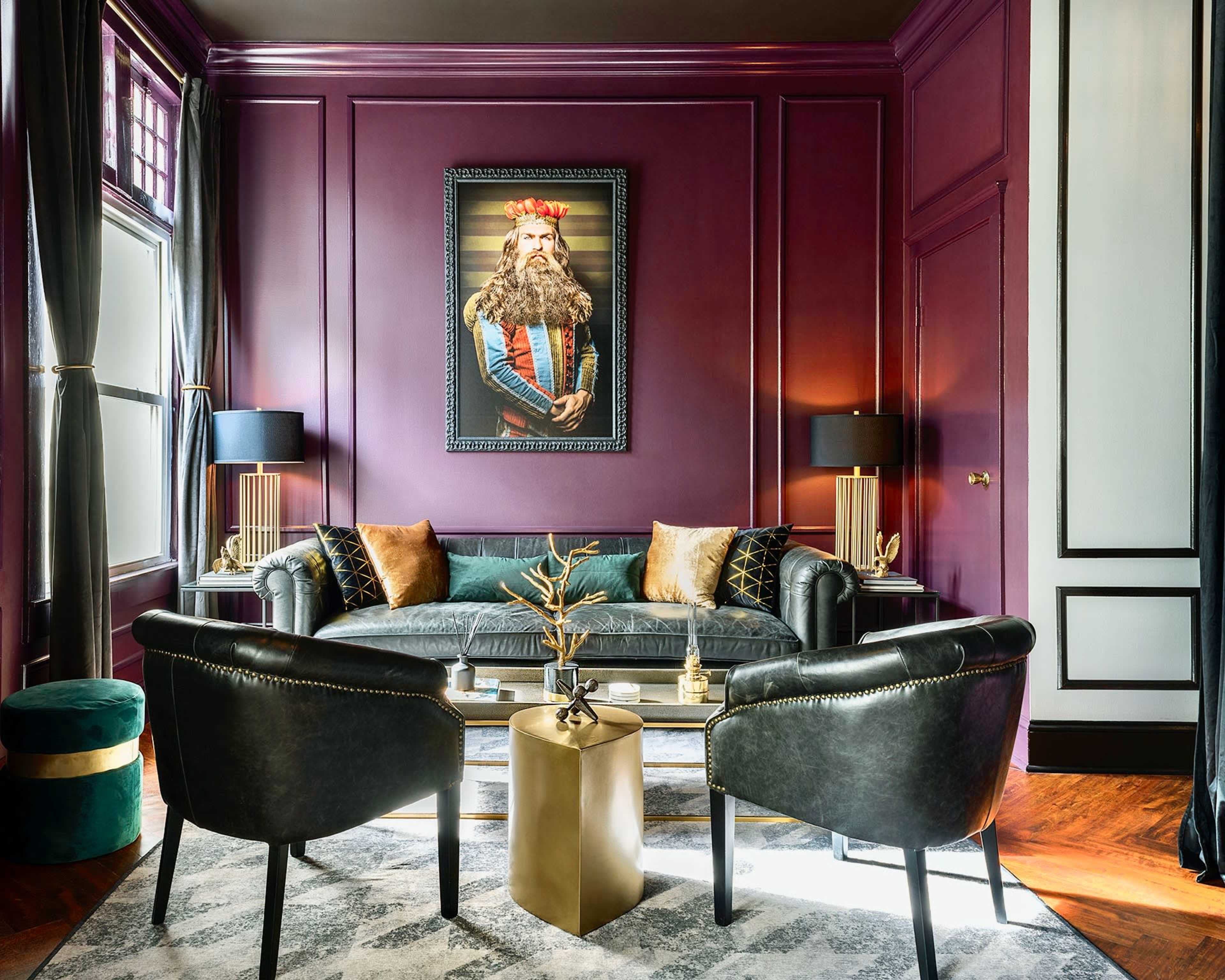 A stylish living room with a deep purple wall, a black leather sofa, two black chairs, and a large portrait of a king hanging above the sofa.