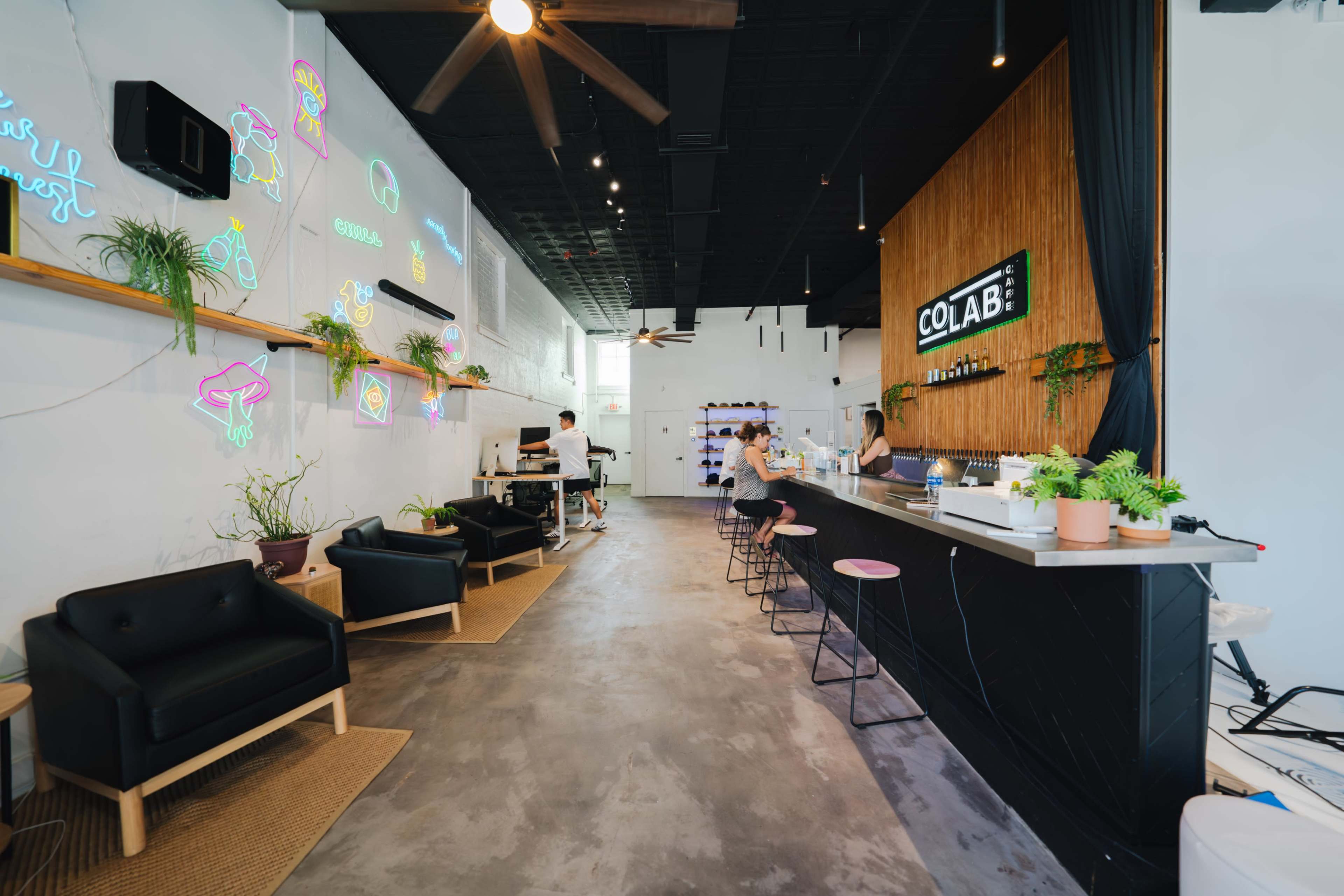 A modern café features a bar with high stools, neon signs on the walls, and a mix of seating areas, including black chairs and greenery.
