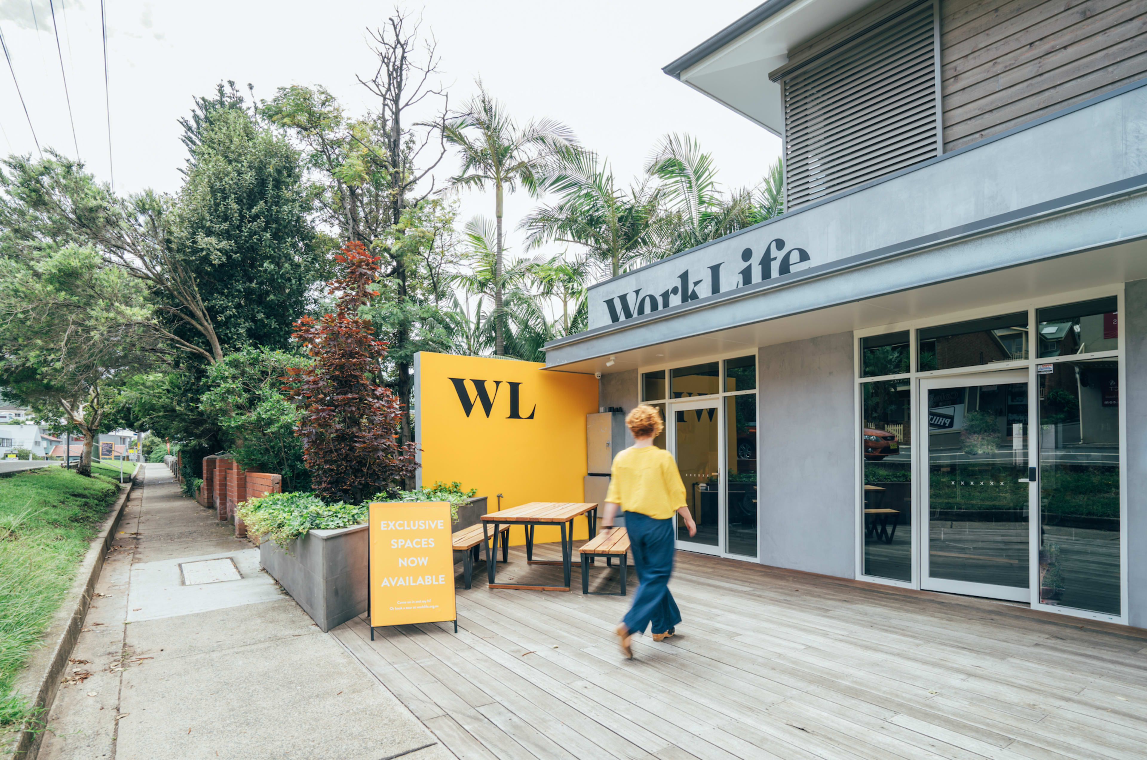 A person walks past a building labeled "Work Life" with a bright yellow wall displaying the letters "WL" and a sign indicating available spaces.