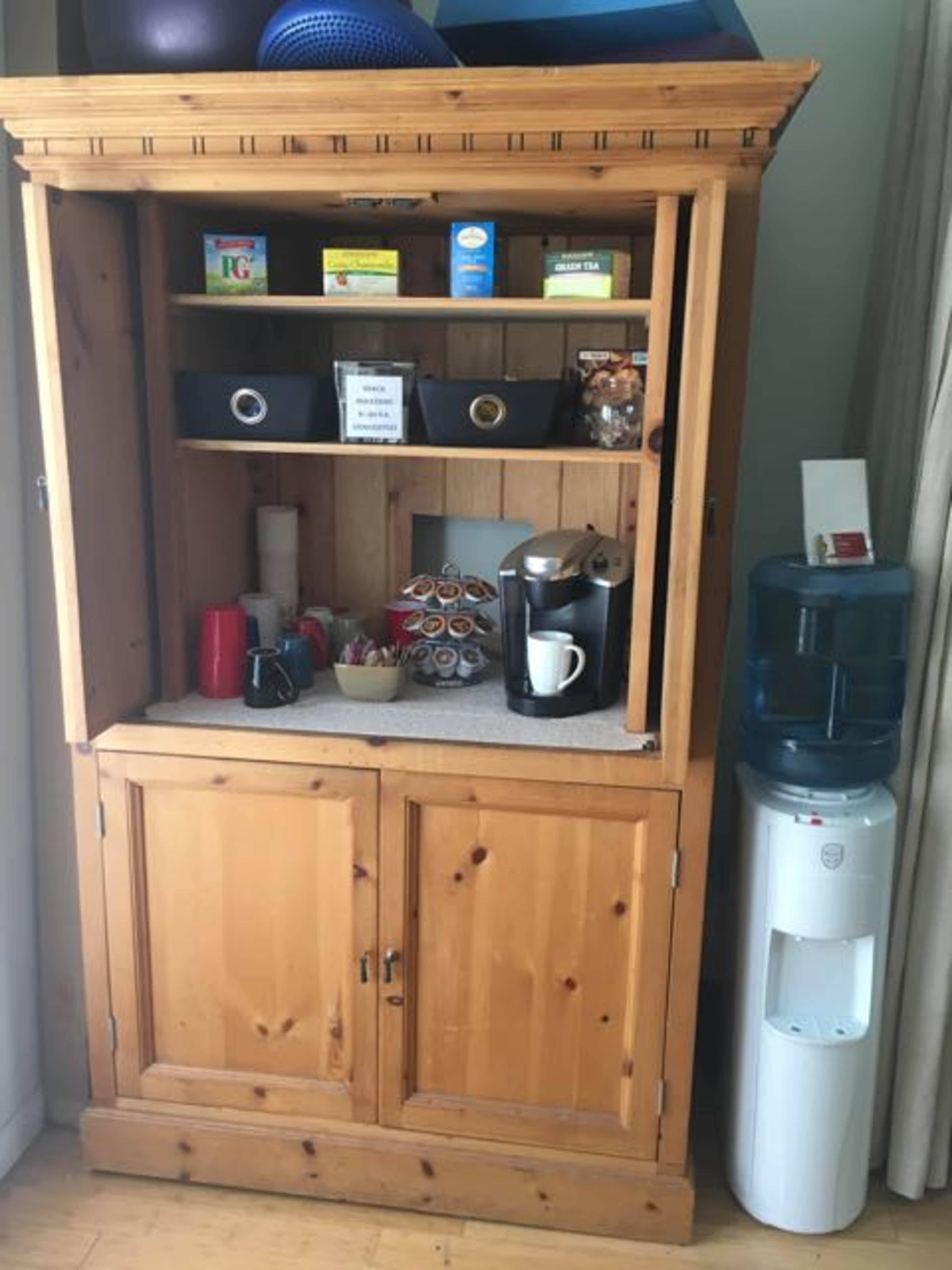 A wooden cabinet with several shelves contains coffee supplies, tea boxes, and a coffee maker, alongside a water cooler on the side.
