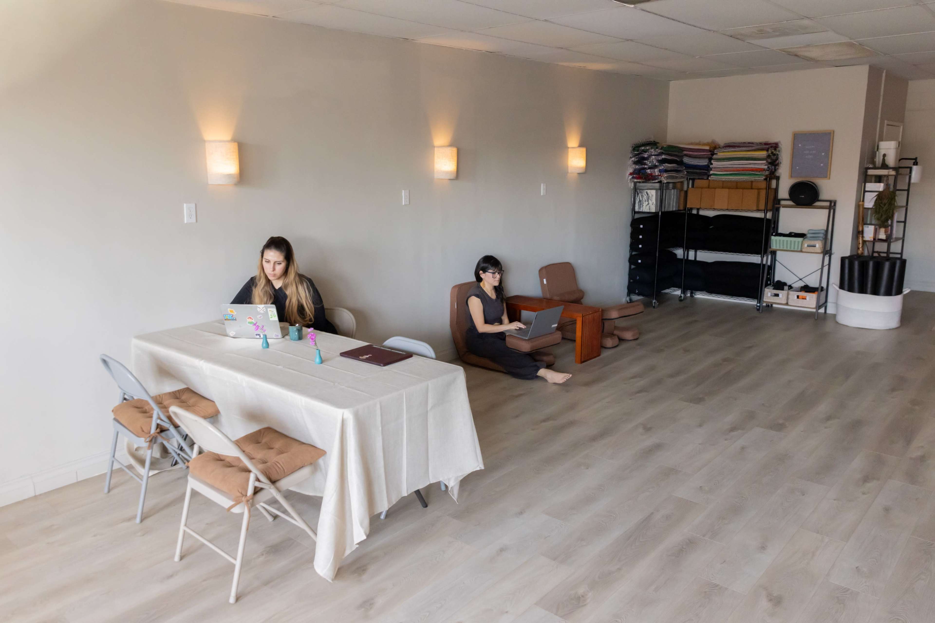 Two individuals are working on laptops in a minimalist, light-colored room with a table and chairs on one side and neatly stacked materials on shelves along the wall.