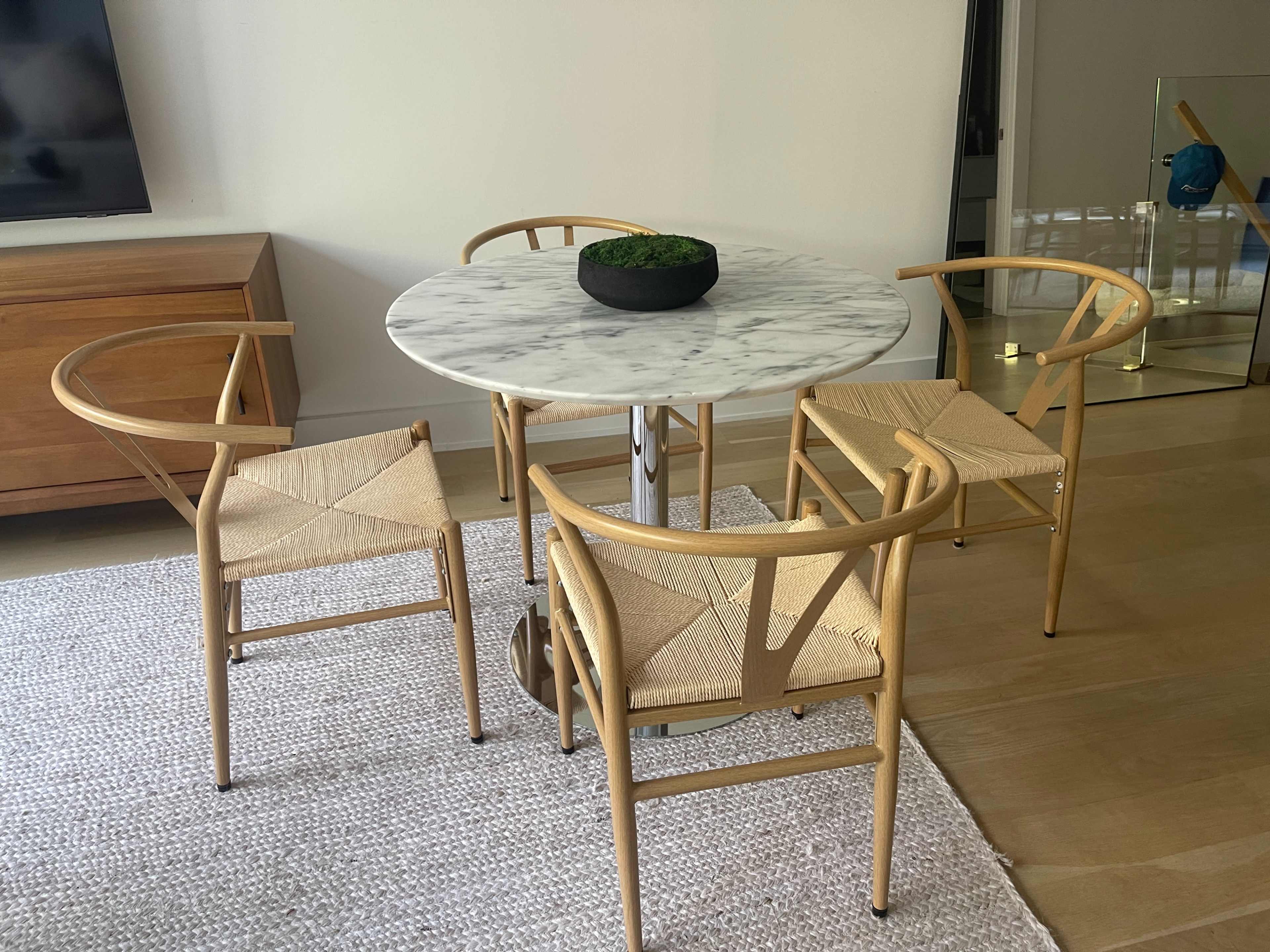 A round marble dining table is surrounded by four wooden chairs with woven seats in a minimalist interior.