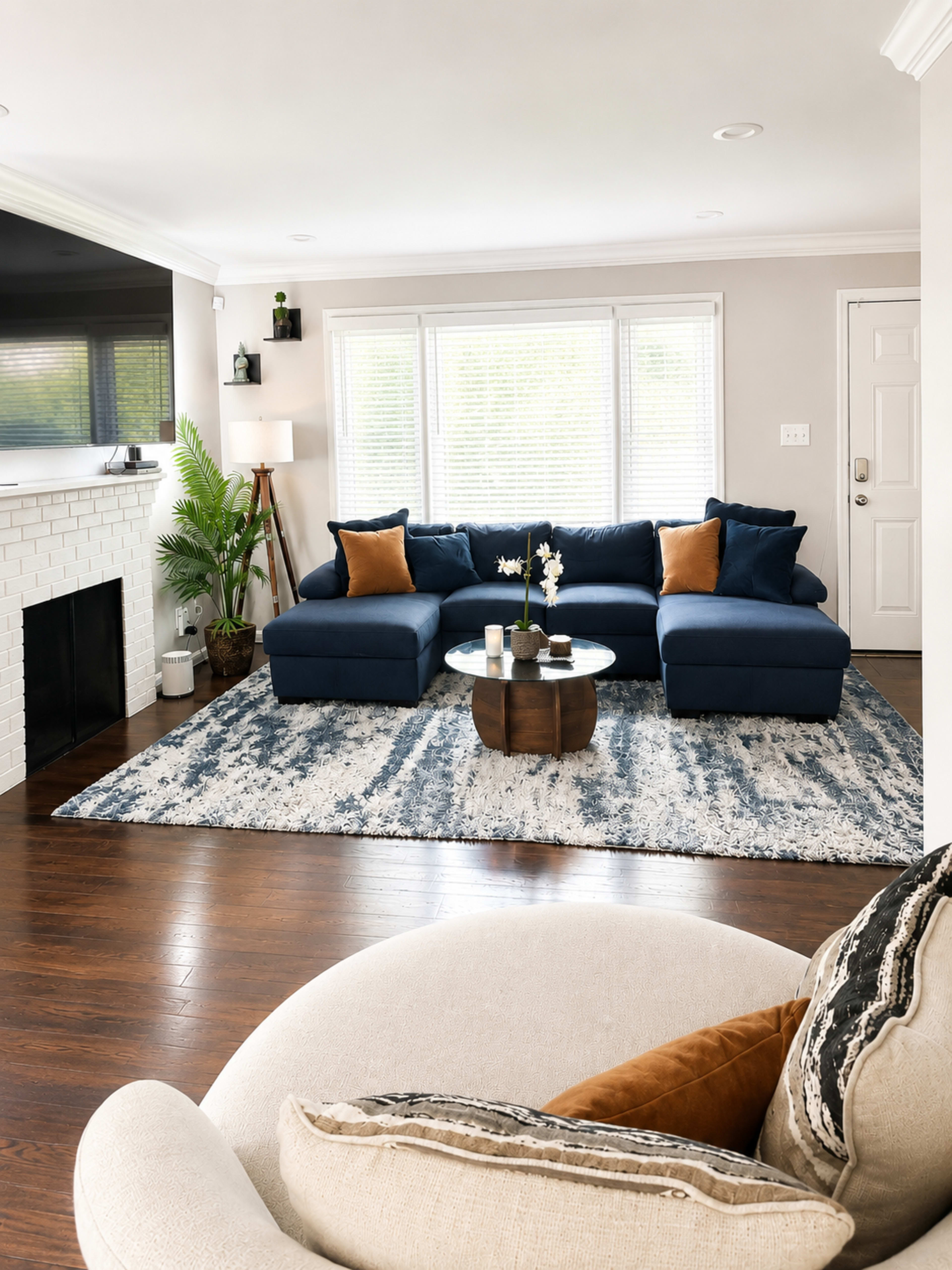 A modern living room features a blue sectional sofa with orange accent pillows, a round coffee table, and a large area rug, surrounded by windows and a white brick fireplace.