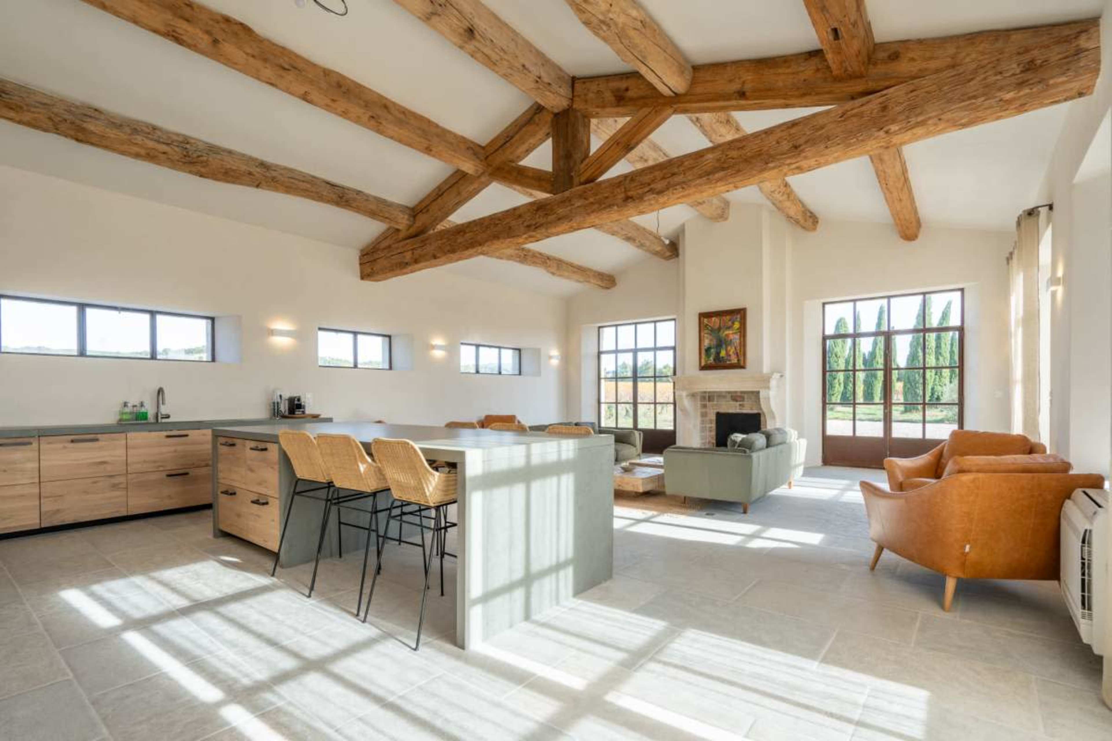 The image shows a modern interior space with a high ceiling supported by wooden beams, featuring a kitchen area, dining seating, and a living area with a fireplace.