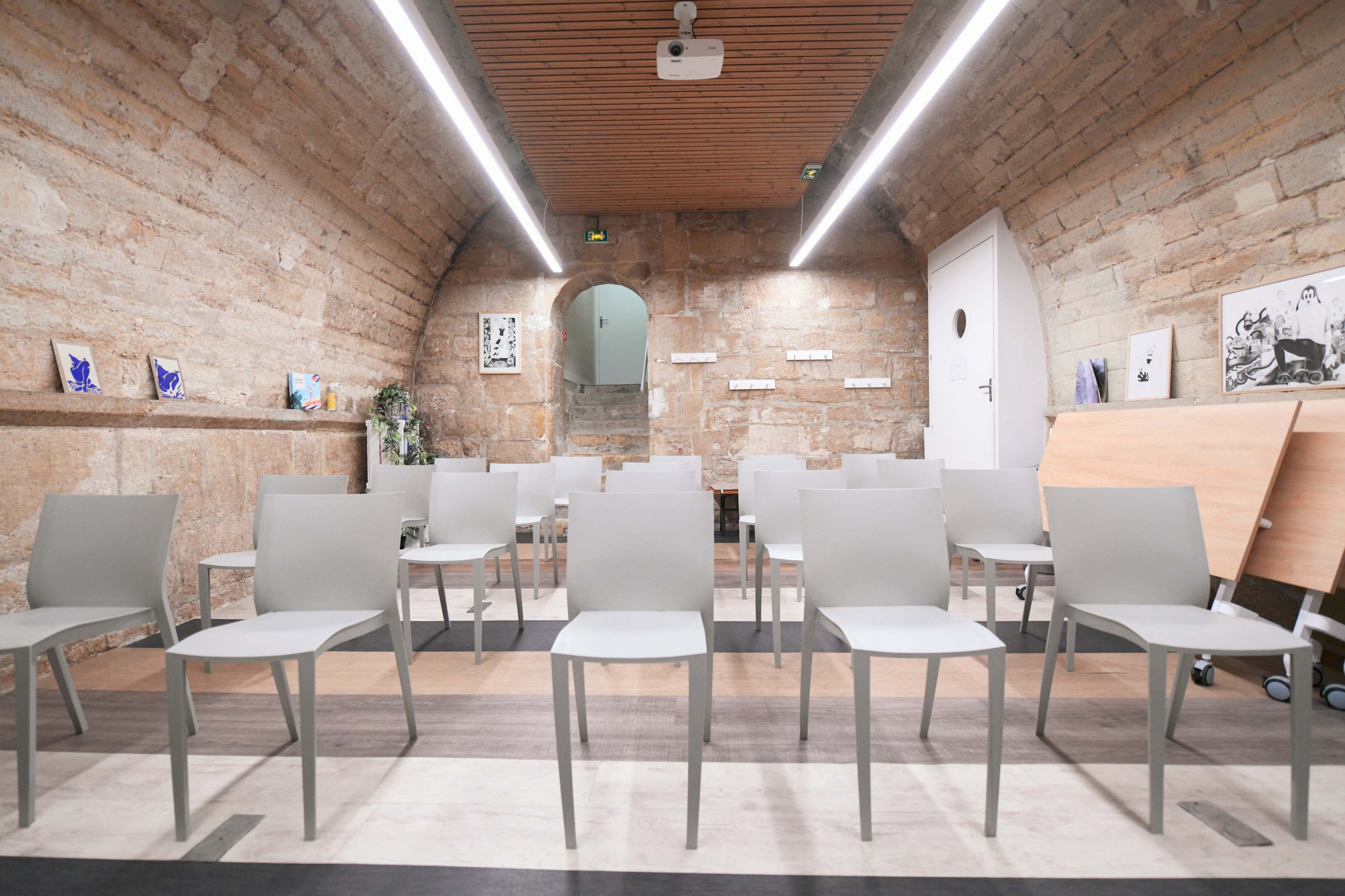 A room with stone walls, featuring rows of gray chairs facing a wall, a projector on the ceiling, and a door visible at the back.