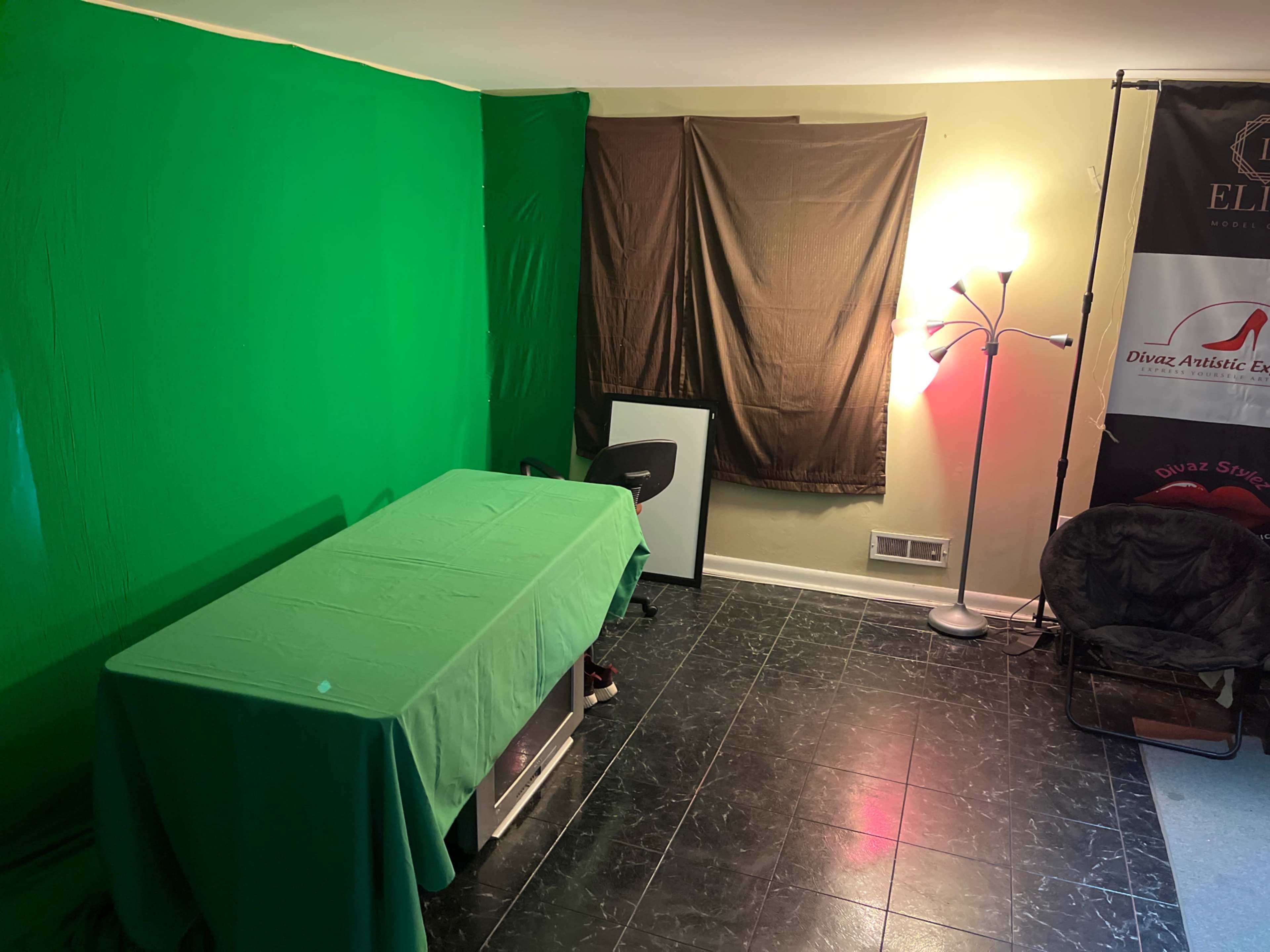 The room contains a green screen, a table covered with a green cloth, a chair, and a lamp beside a dark curtain and a banner.
