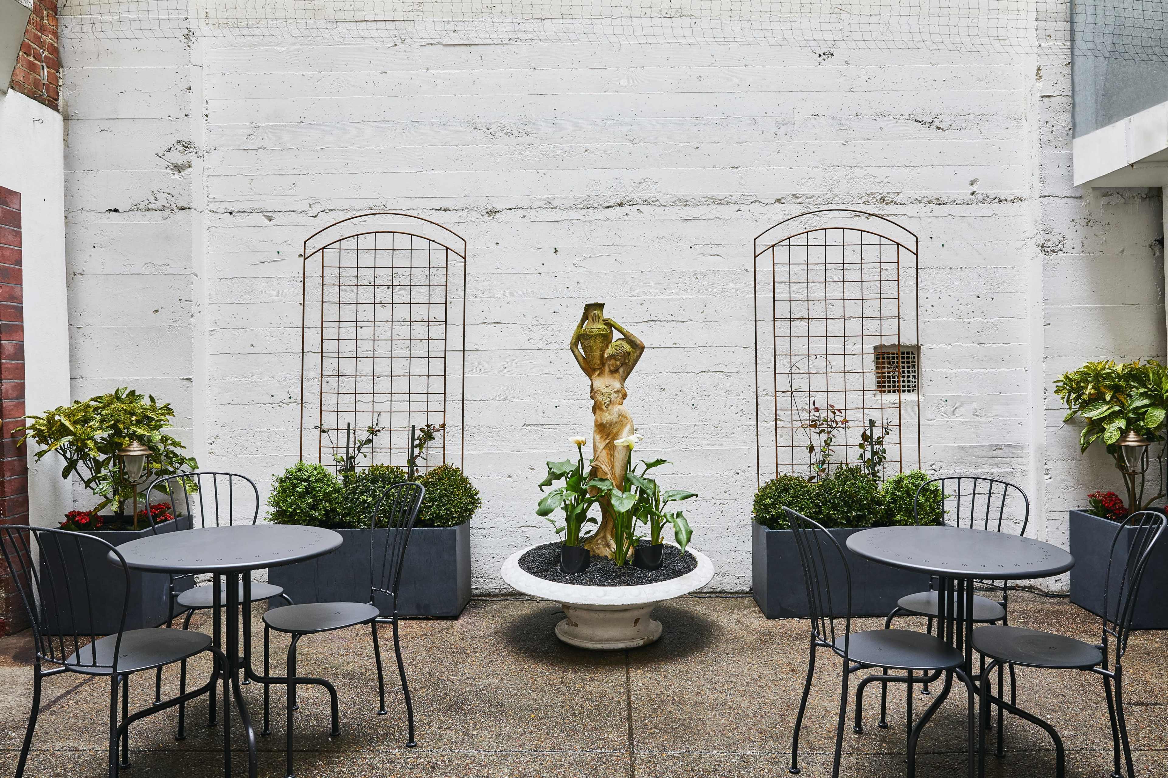 A courtyard with four round, black metal tables and chairs surrounding a central golden statue in a planter, flanked by trellises and greenery against a textured concrete wall.
