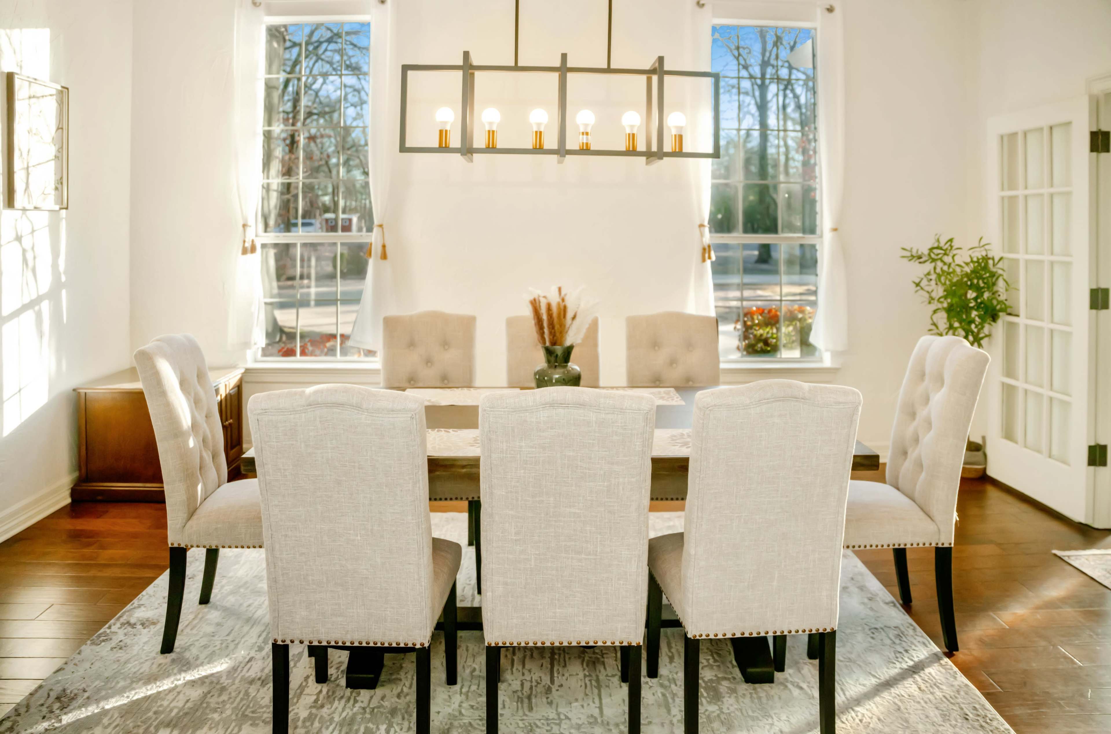 A dining room features a rectangular table surrounded by six upholstered chairs, with large windows and a hanging light fixture above.