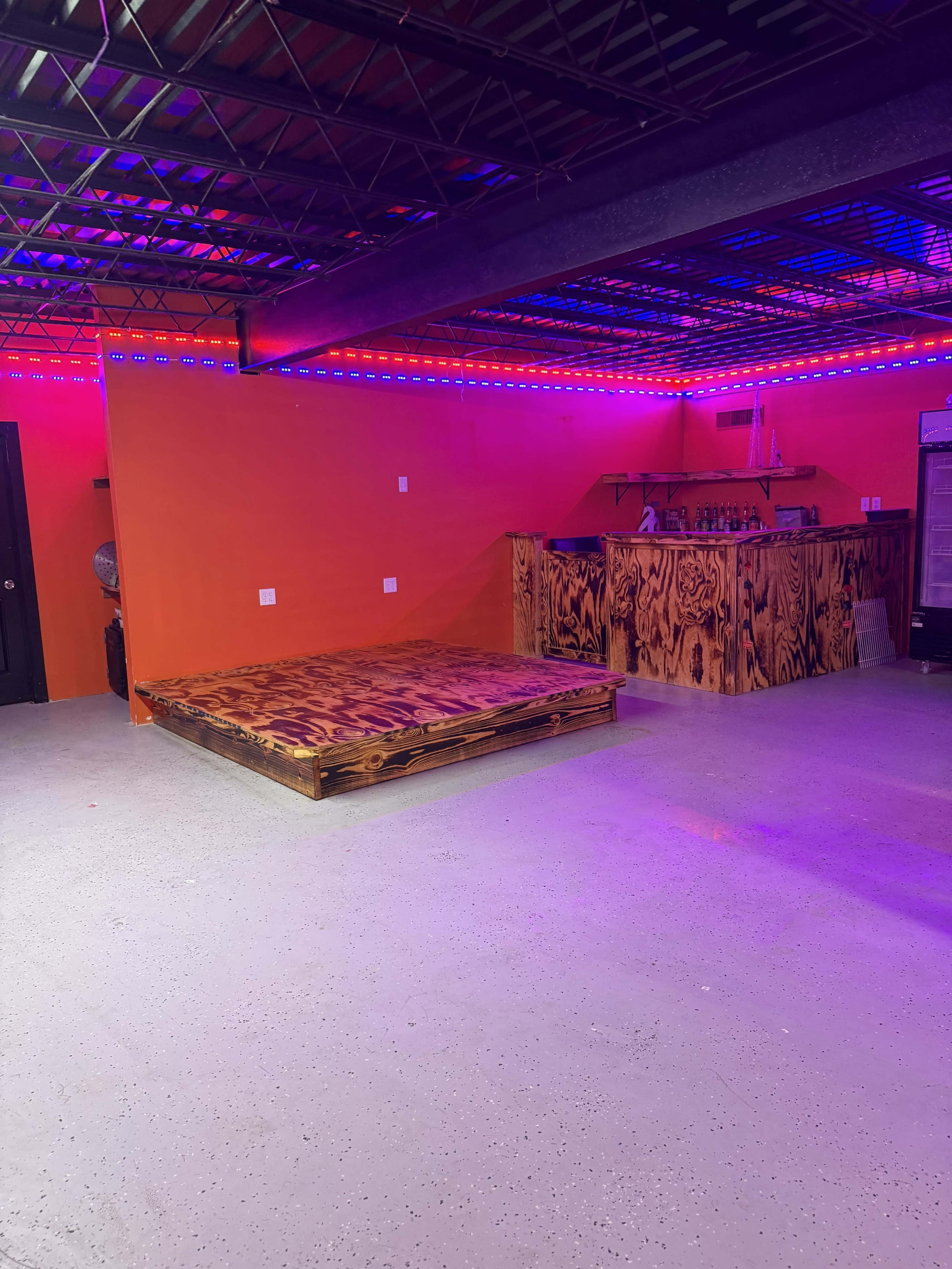 The image shows a sparsely furnished room with a wooden stage platform, a bar area in the back, and colorful lighting along the ceiling.