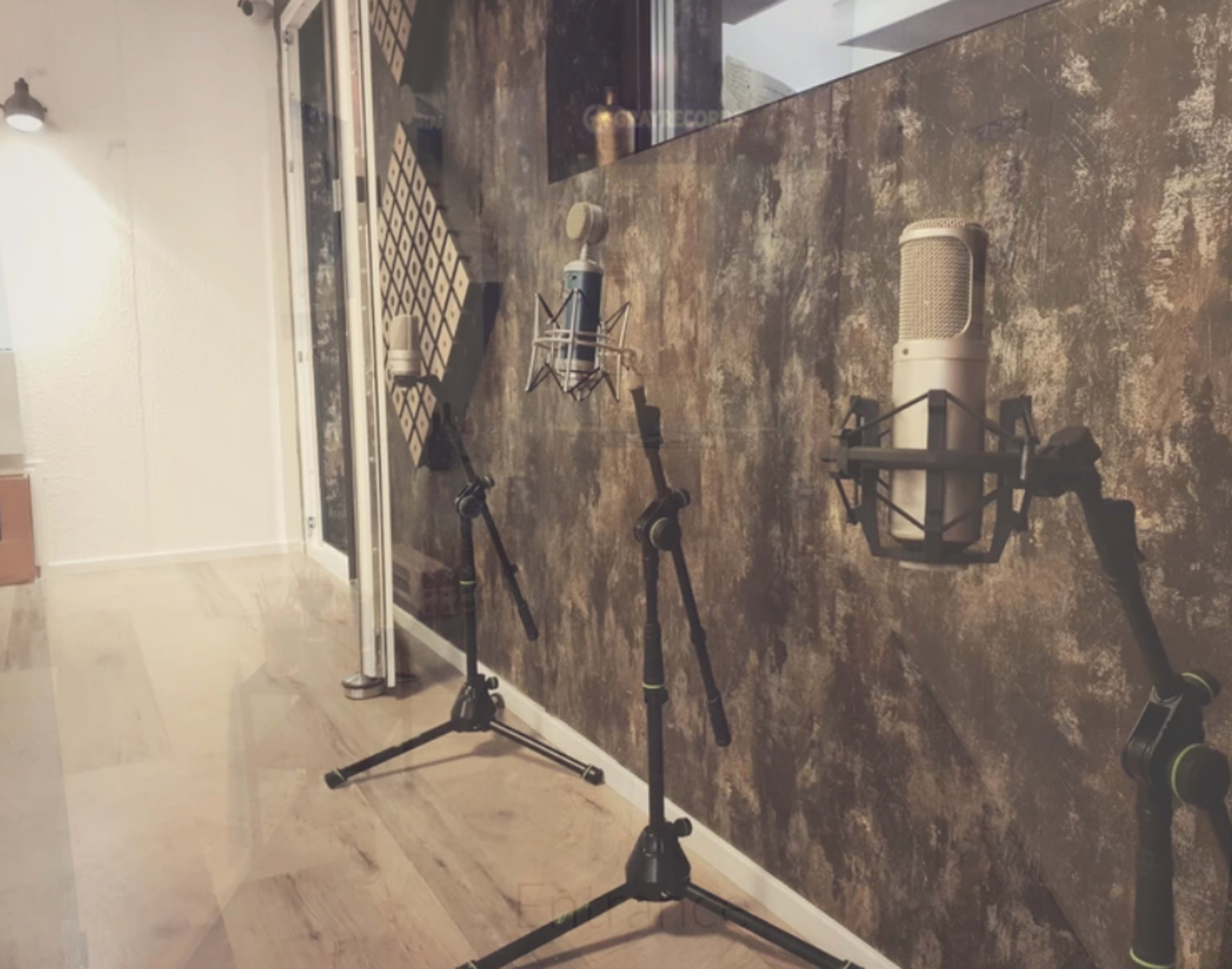The image shows three microphones mounted on stands in a room with a textured wall and wooden flooring.