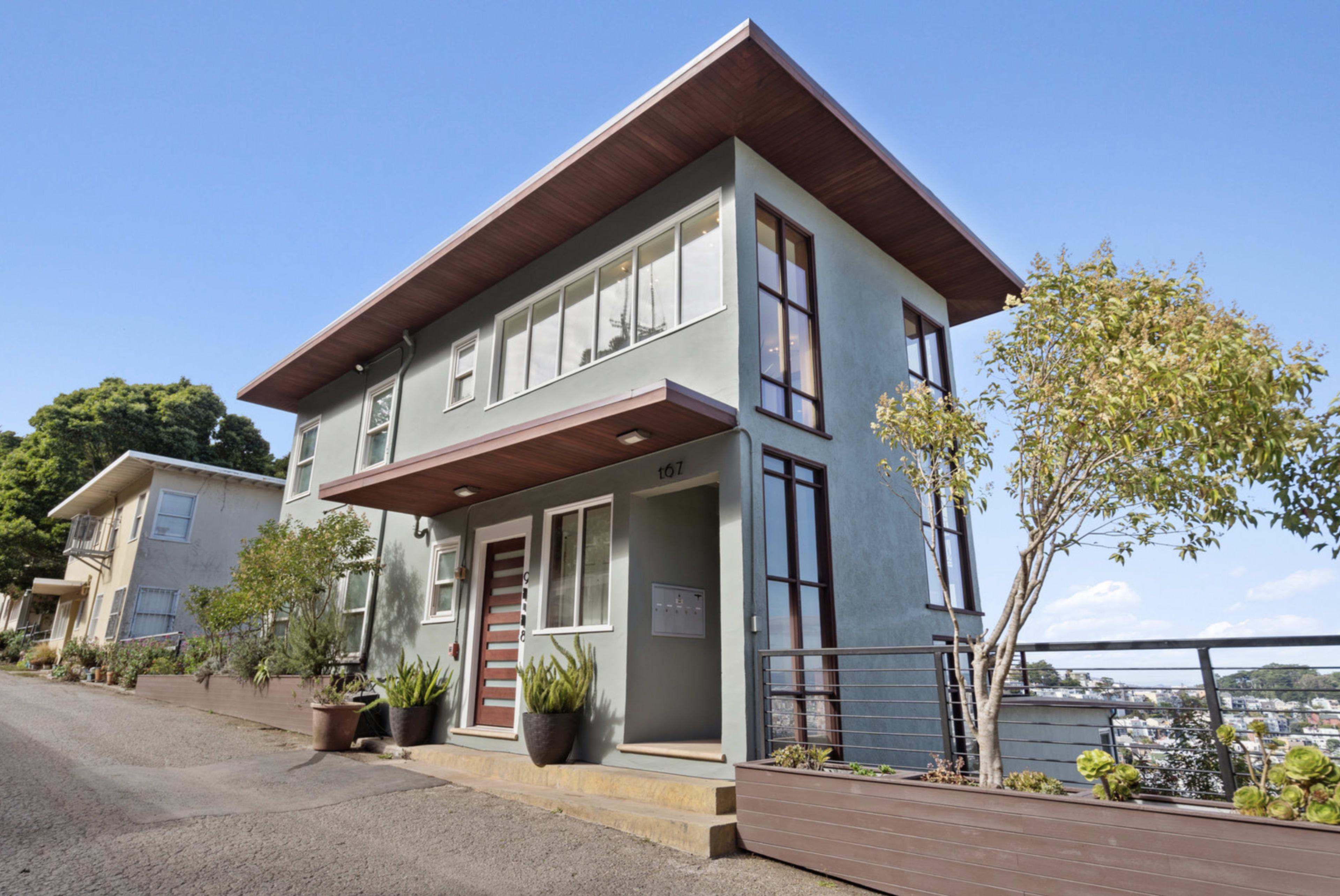 The image shows a modern, two-story house with a flat roof, large windows, and a landscaped entrance located on a hillside.