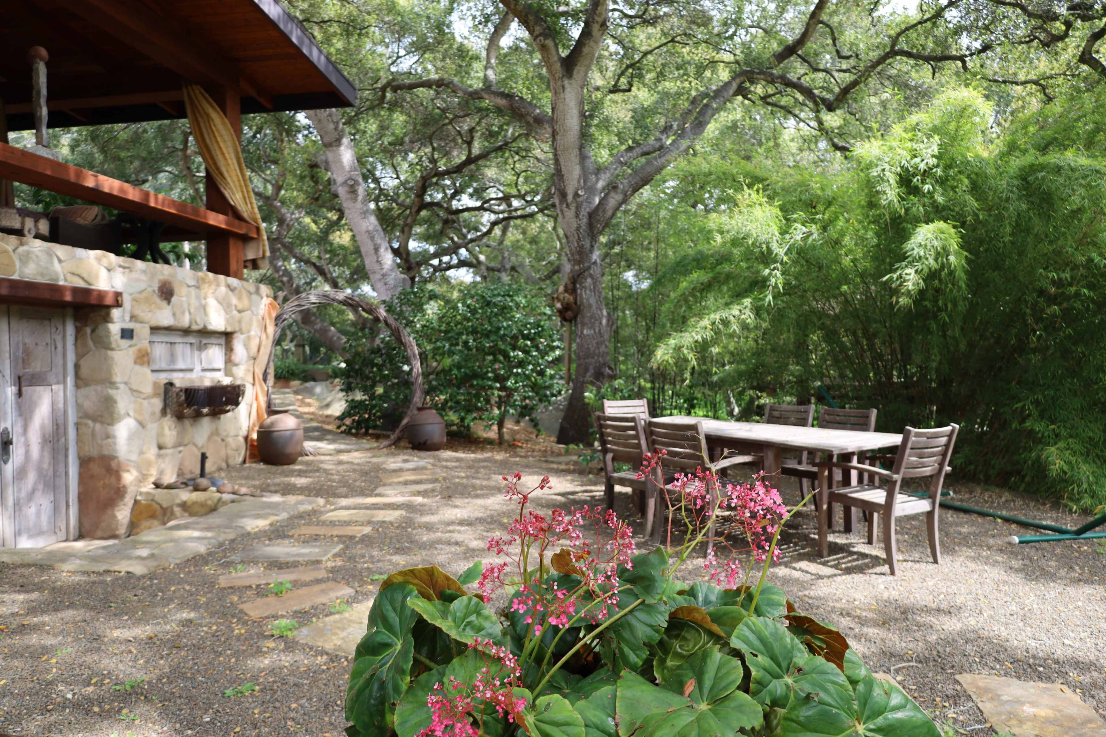 A stone patio features a wooden table and chairs surrounded by greenery and flowering plants.
