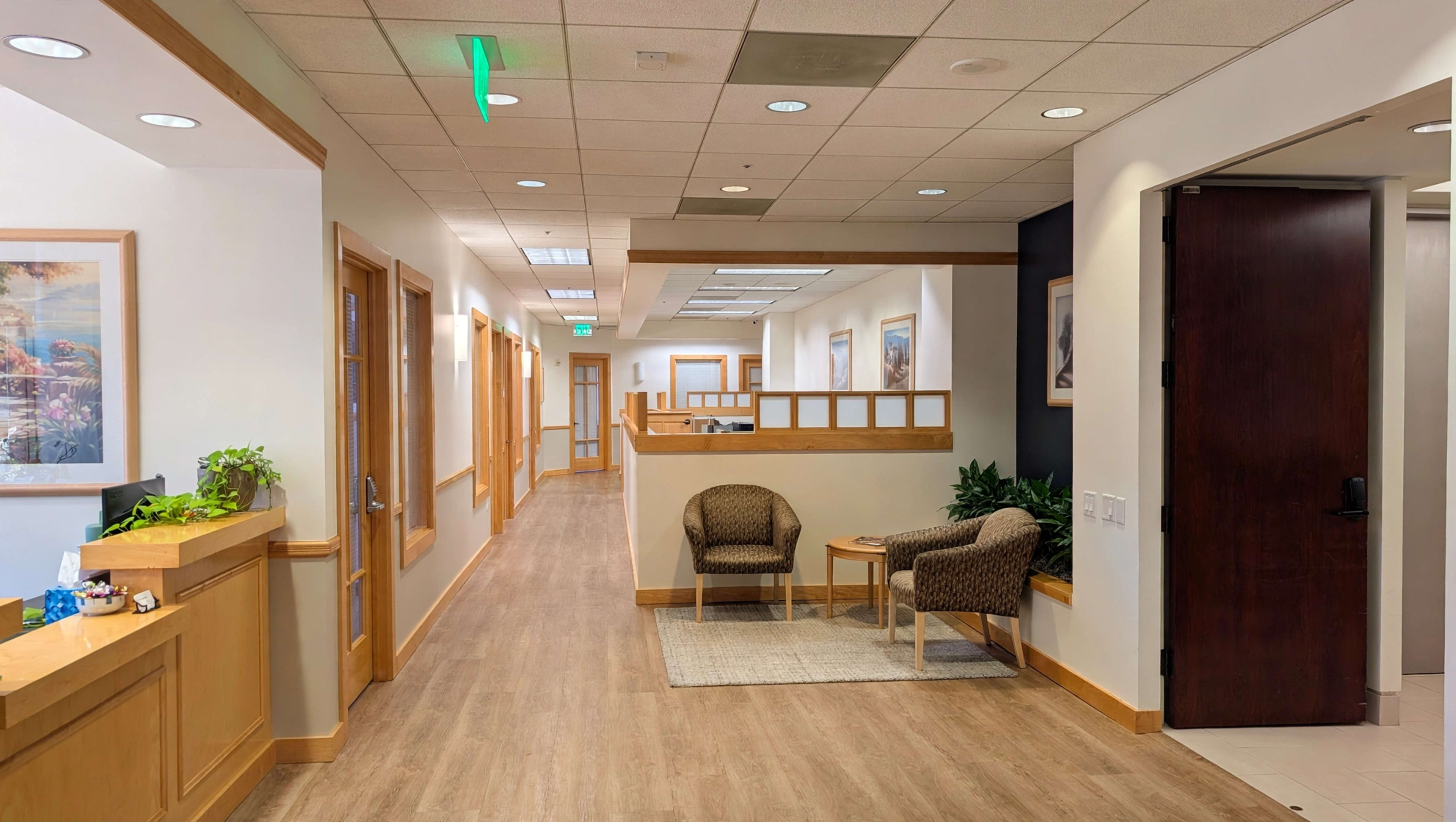 The image shows a modern interior hallway of a building with a reception area, two seating chairs, and wooden accents.