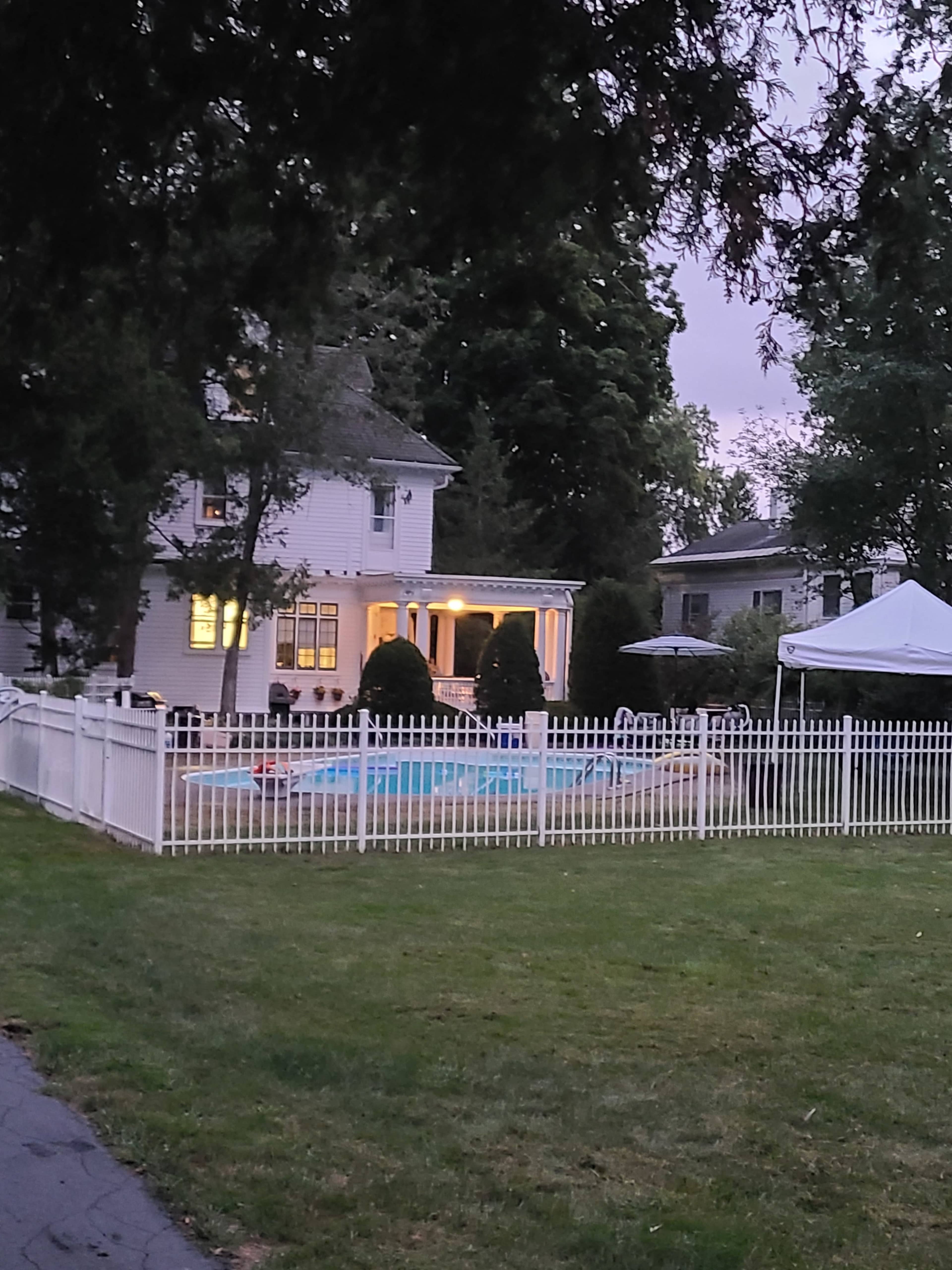 A white house with a porch overlooks a fenced-in pool area, surrounded by trees and a tent on the lawn.