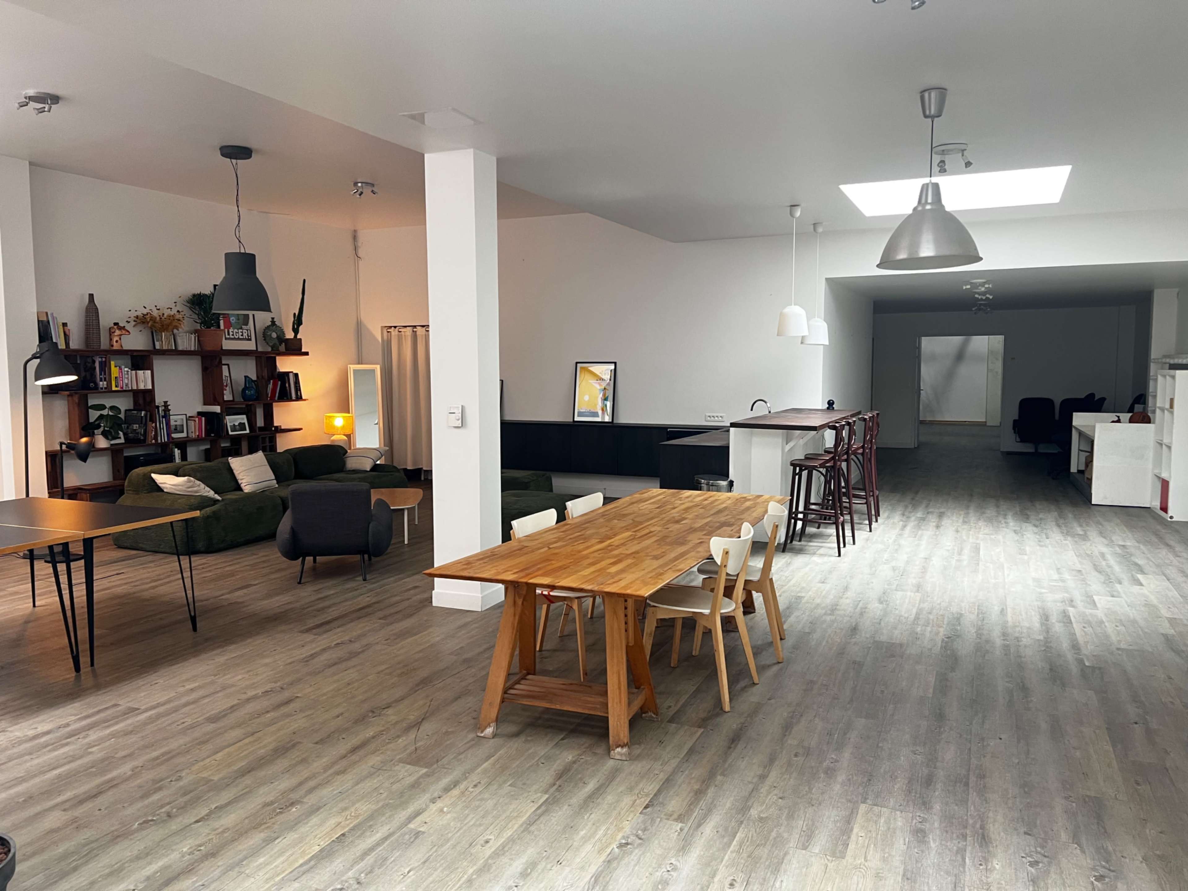 The image shows a spacious, modern interior with a dining table and chairs, a living area with green seating, and a kitchenette visible in the background.