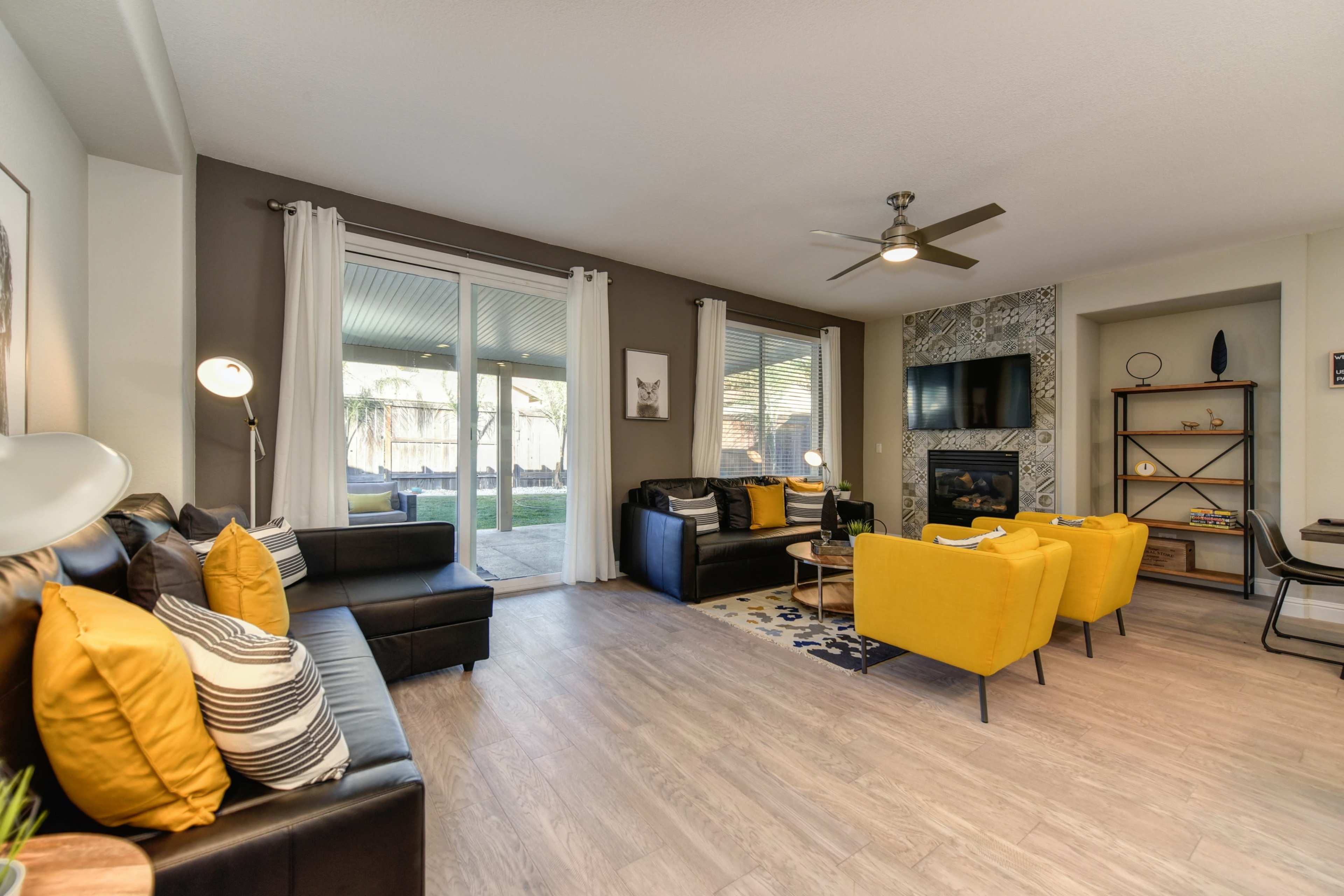 The living room features black couches, yellow armchairs, a television, and large windows overlooking a patio.
