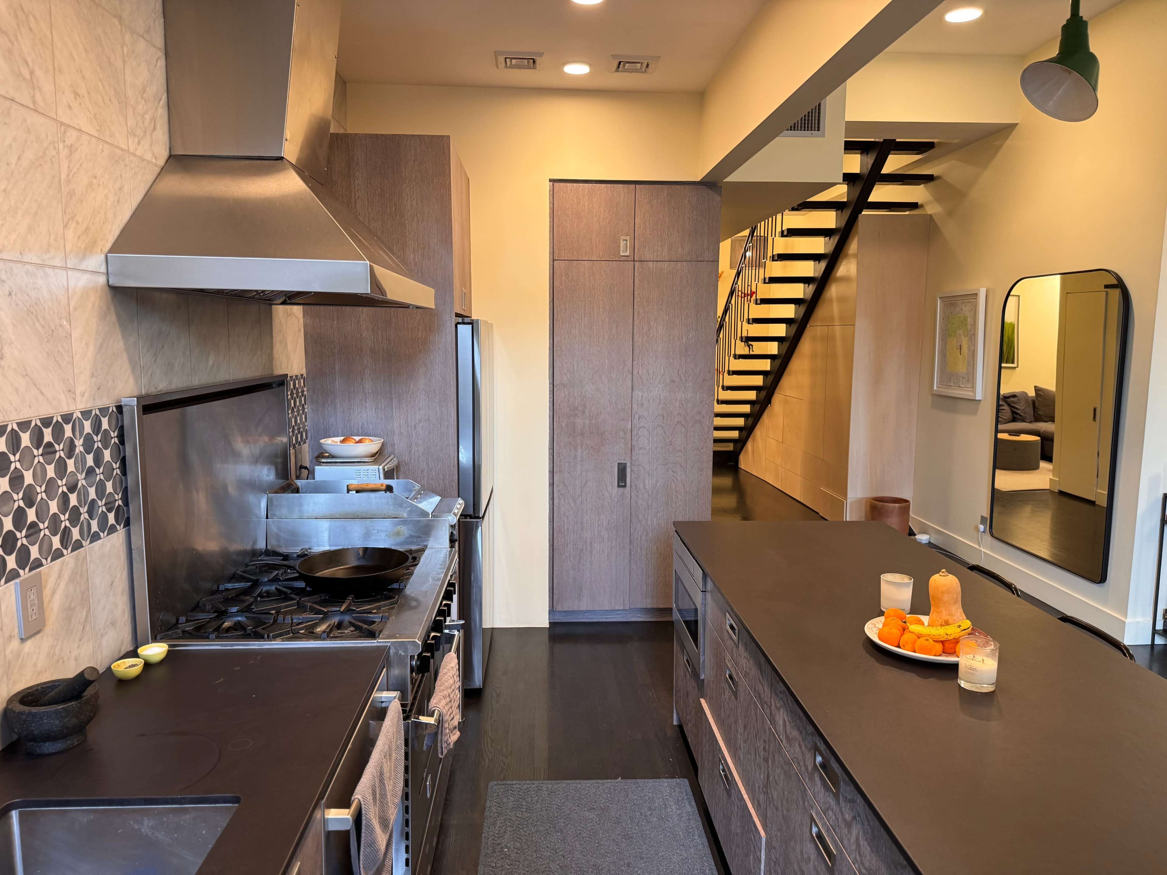 A modern kitchen features stainless steel appliances, a spacious island with fruit, and a staircase leading to another level in the background.