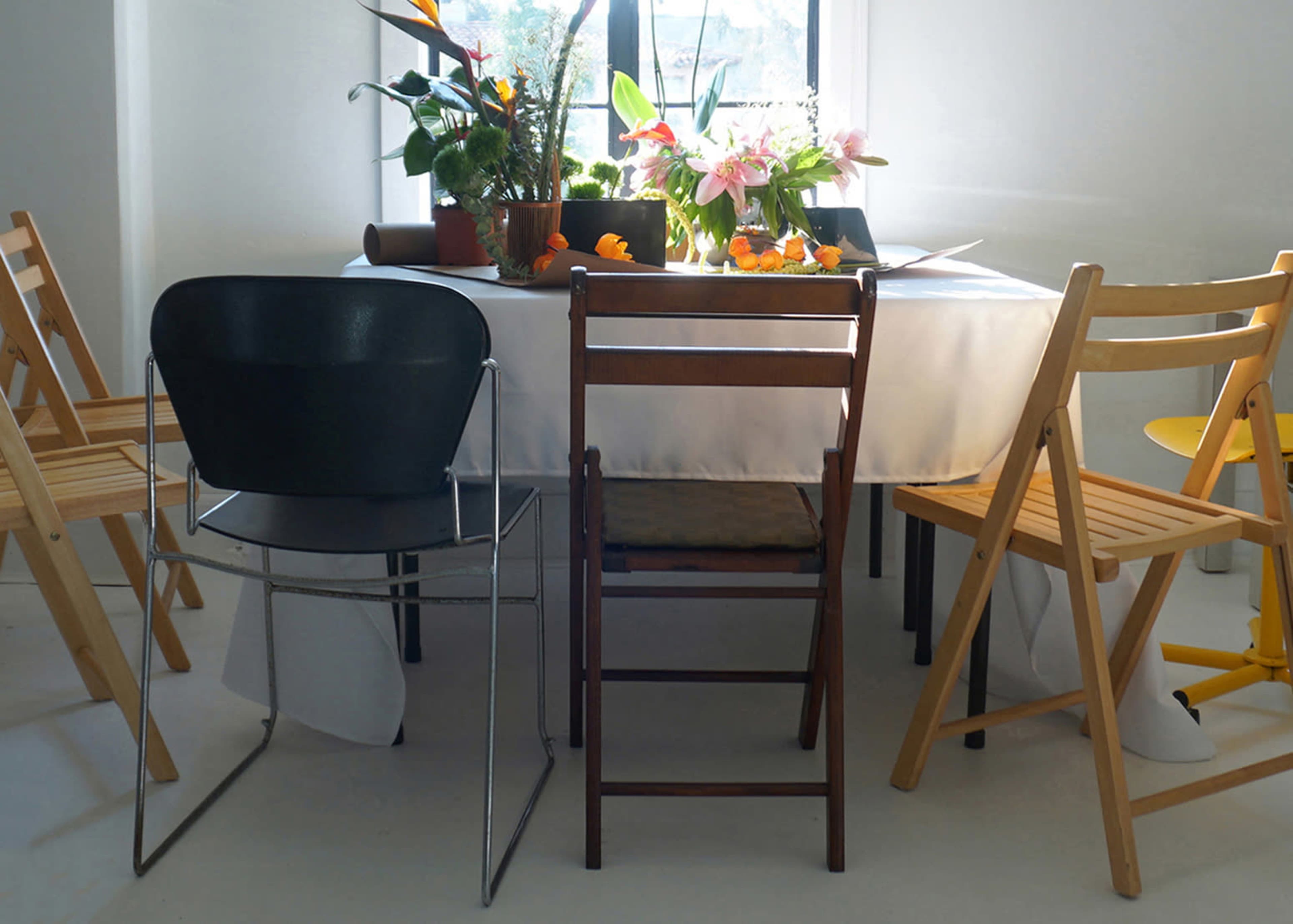 A round table with a white tablecloth is surrounded by various types of chairs, and there are potted plants and flowers on the table.