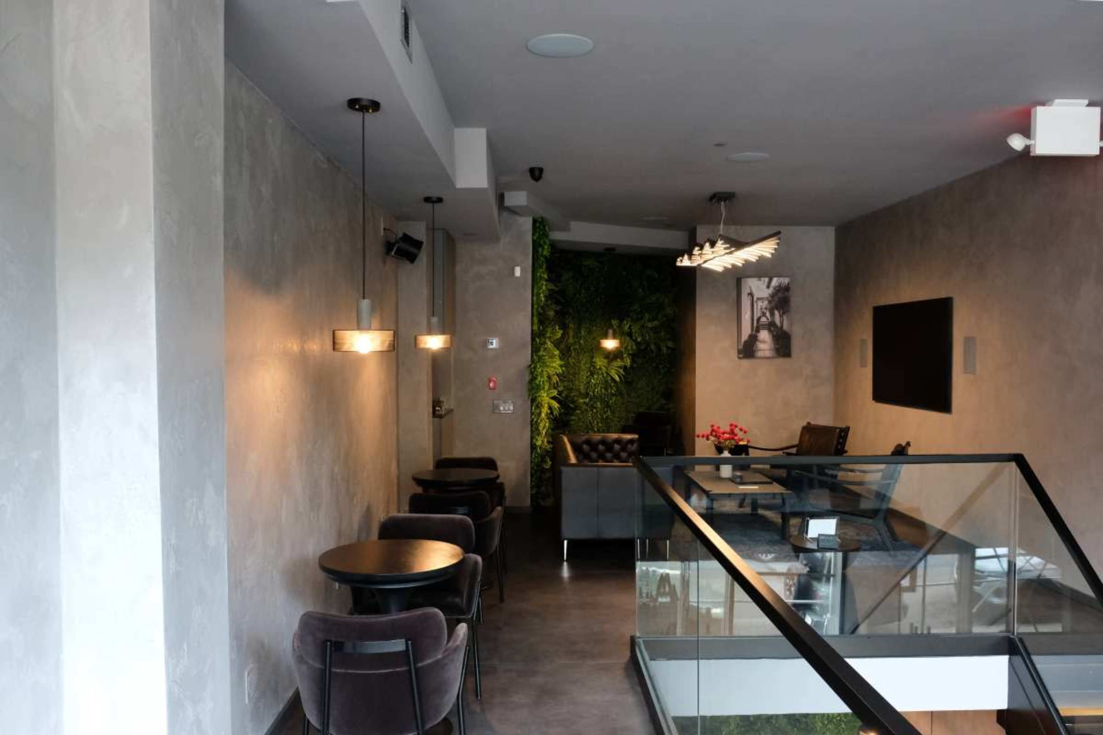 The image shows a modern interior of a cafe or lounge area with dark furniture, green wall plants, and soft lighting.