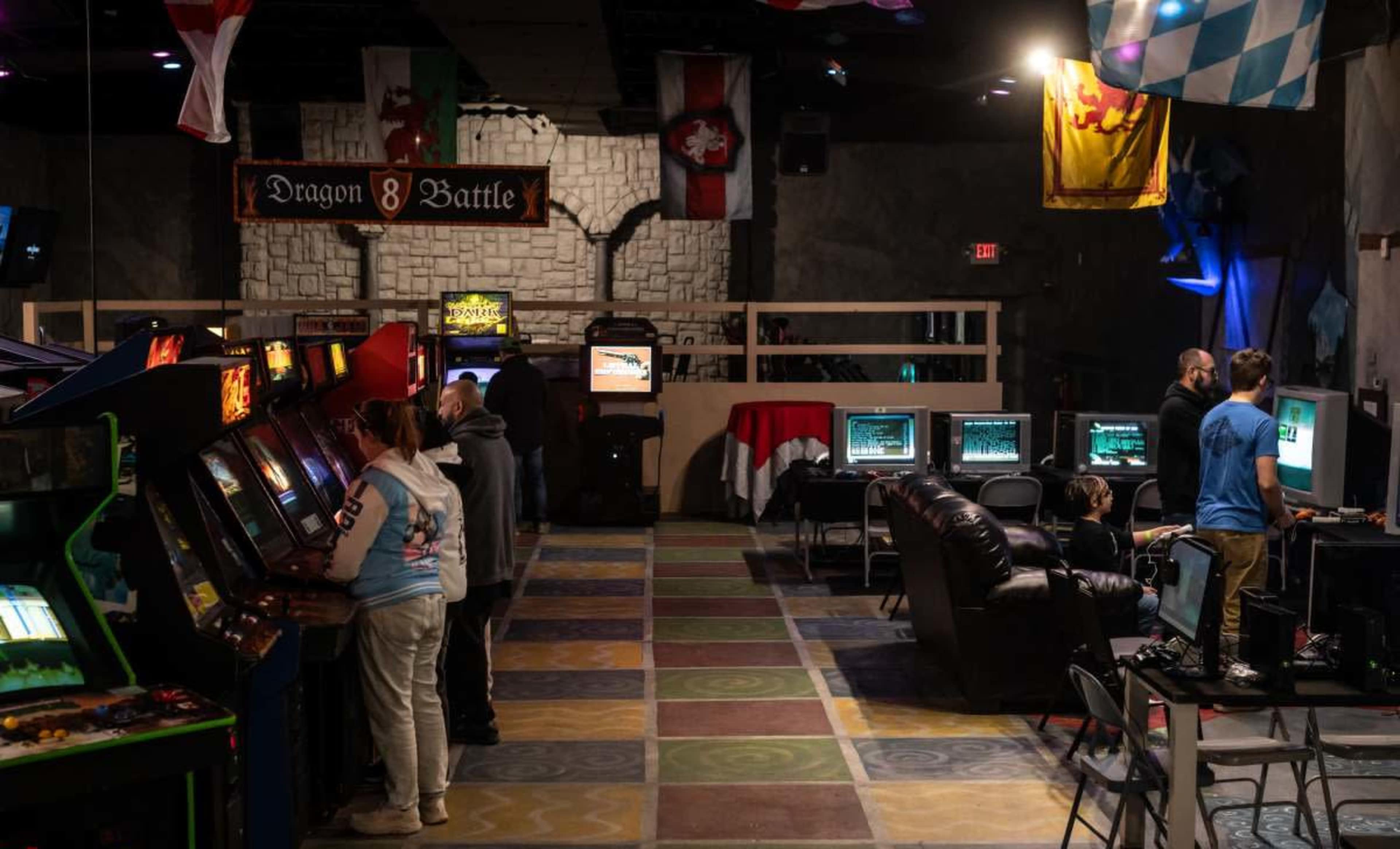 The image shows an arcade with various vintage video game machines and several people engaged with the games in a colorful, checkered floor environment.