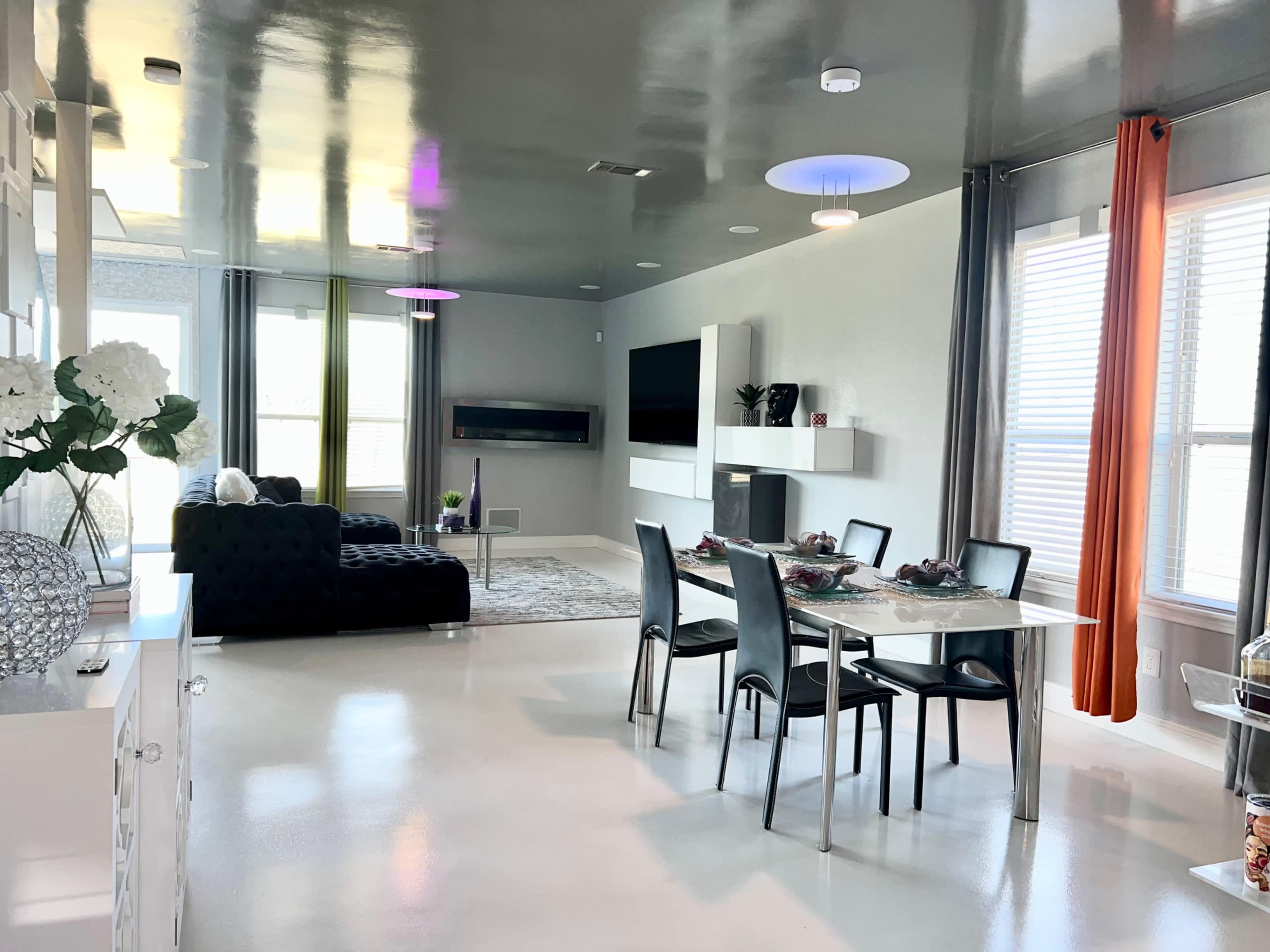 A modern living space features a dining area with a glass table and chairs, a black sofa, and a large window with sheer curtains.