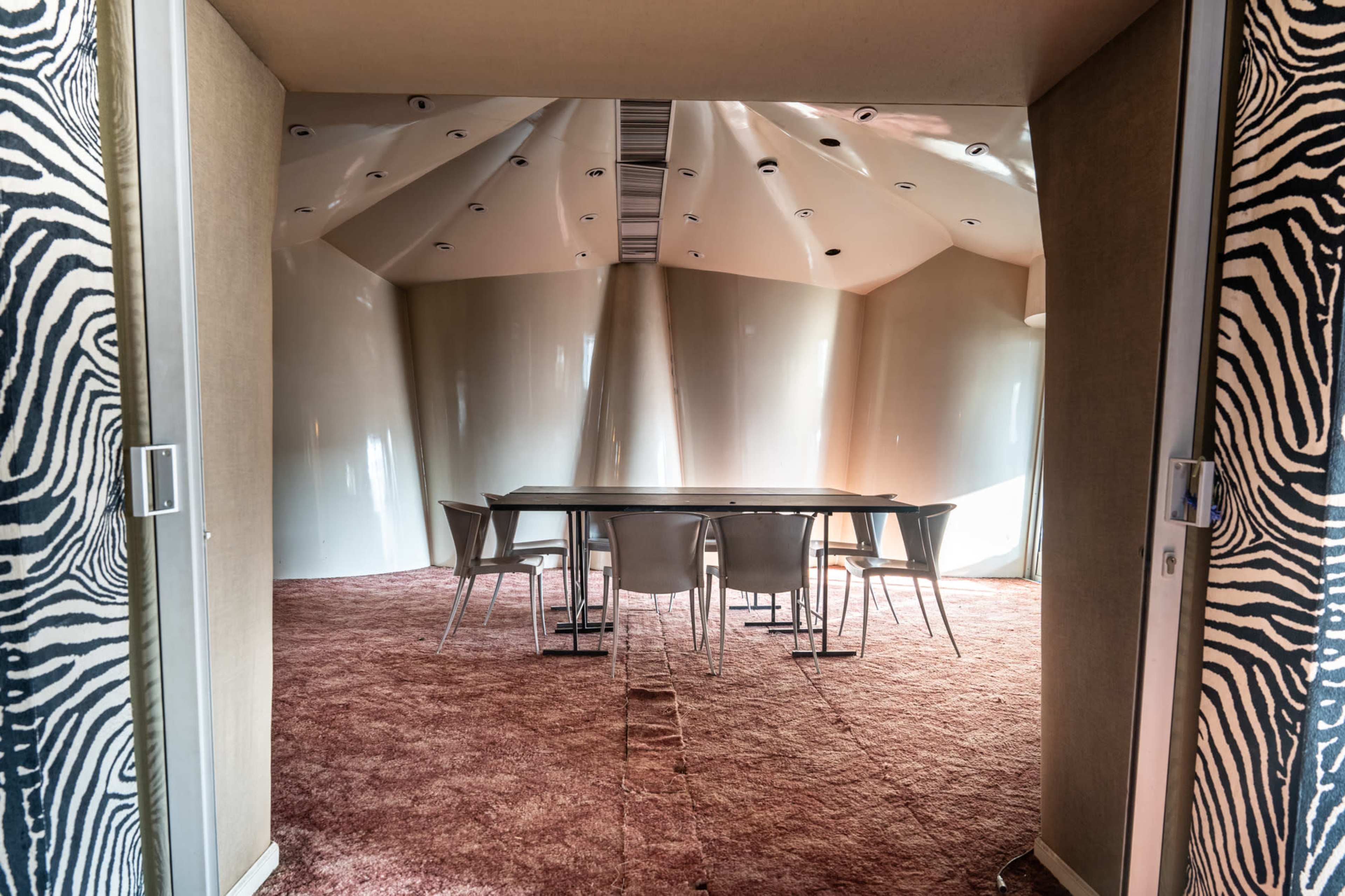 The image shows a modern, minimalist meeting room with a large table and several chairs, featuring curved walls and a textured carpet.