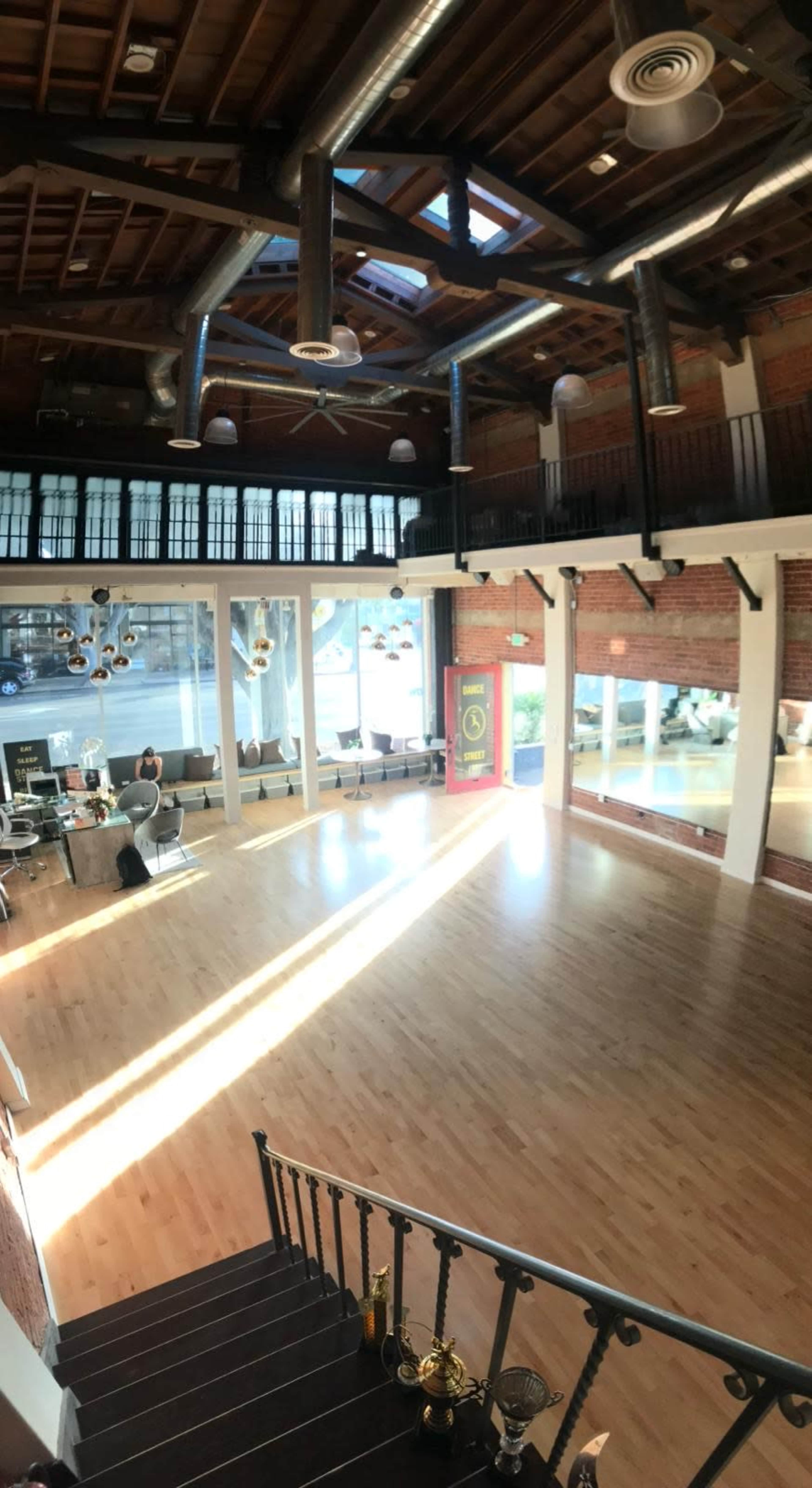 The image shows a spacious, well-lit room with wooden floors, large windows, and exposed brick walls, designed for gatherings or events.