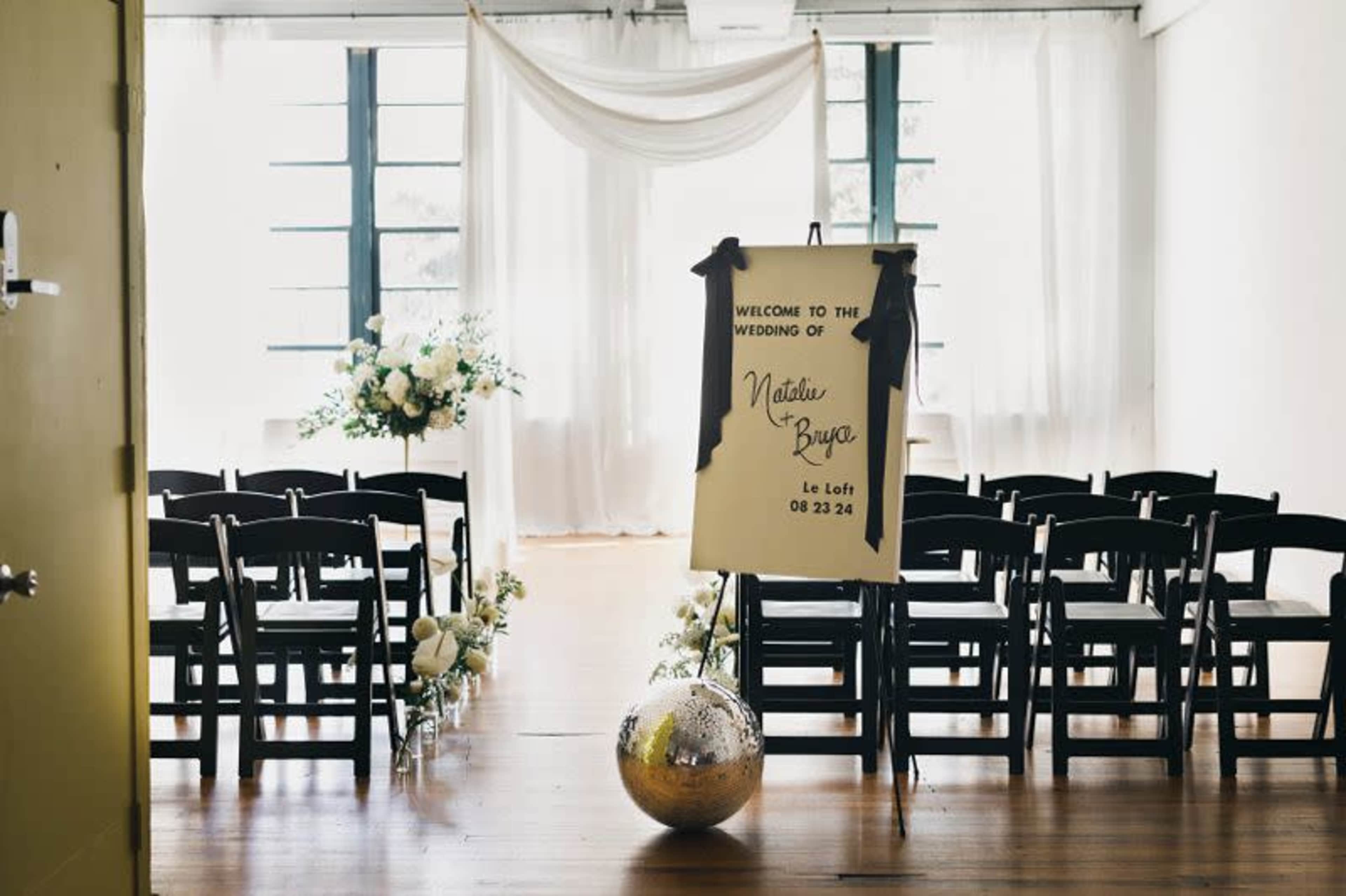 The image shows a wedding setup in a venue with rows of black chairs facing a decorative backdrop, featuring floral arrangements and a sign welcoming guests.