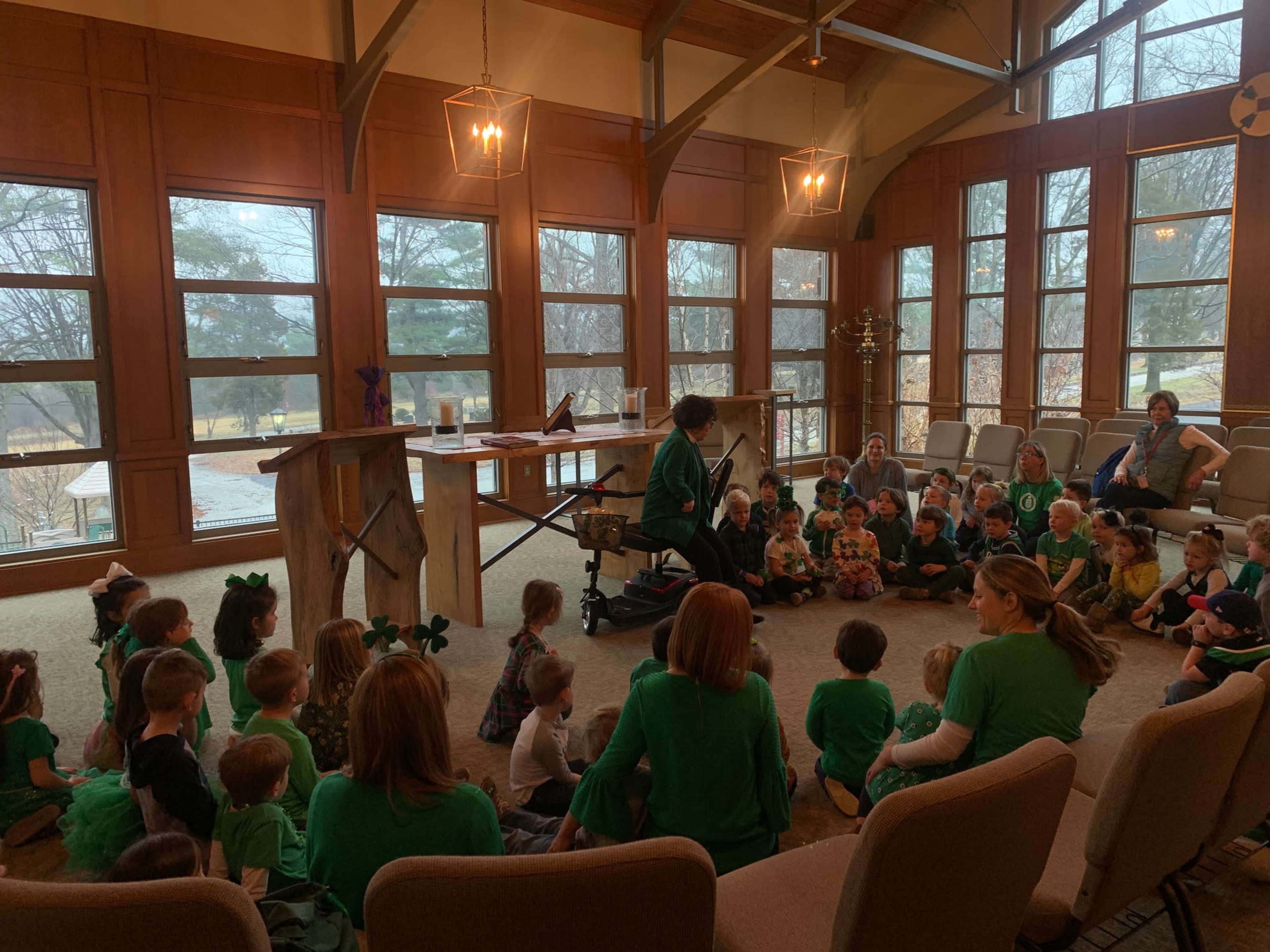 Young children in green outfits sit in a circle on the floor while an adult leads an activity in a room with large windows.