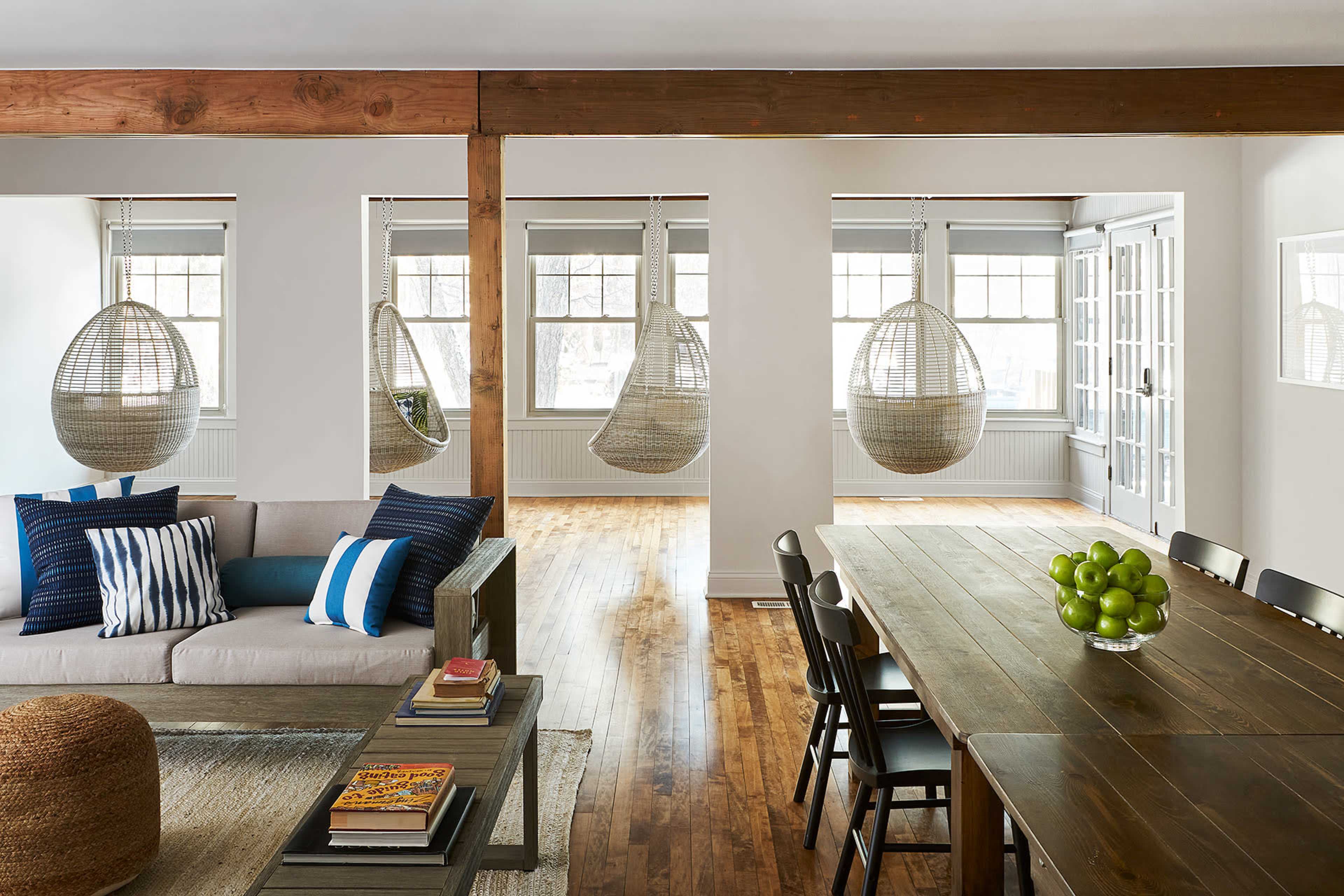 A modern, airy living space featuring a couch with decorative pillows, a dining table with chairs, and hanging wicker chairs near large windows.