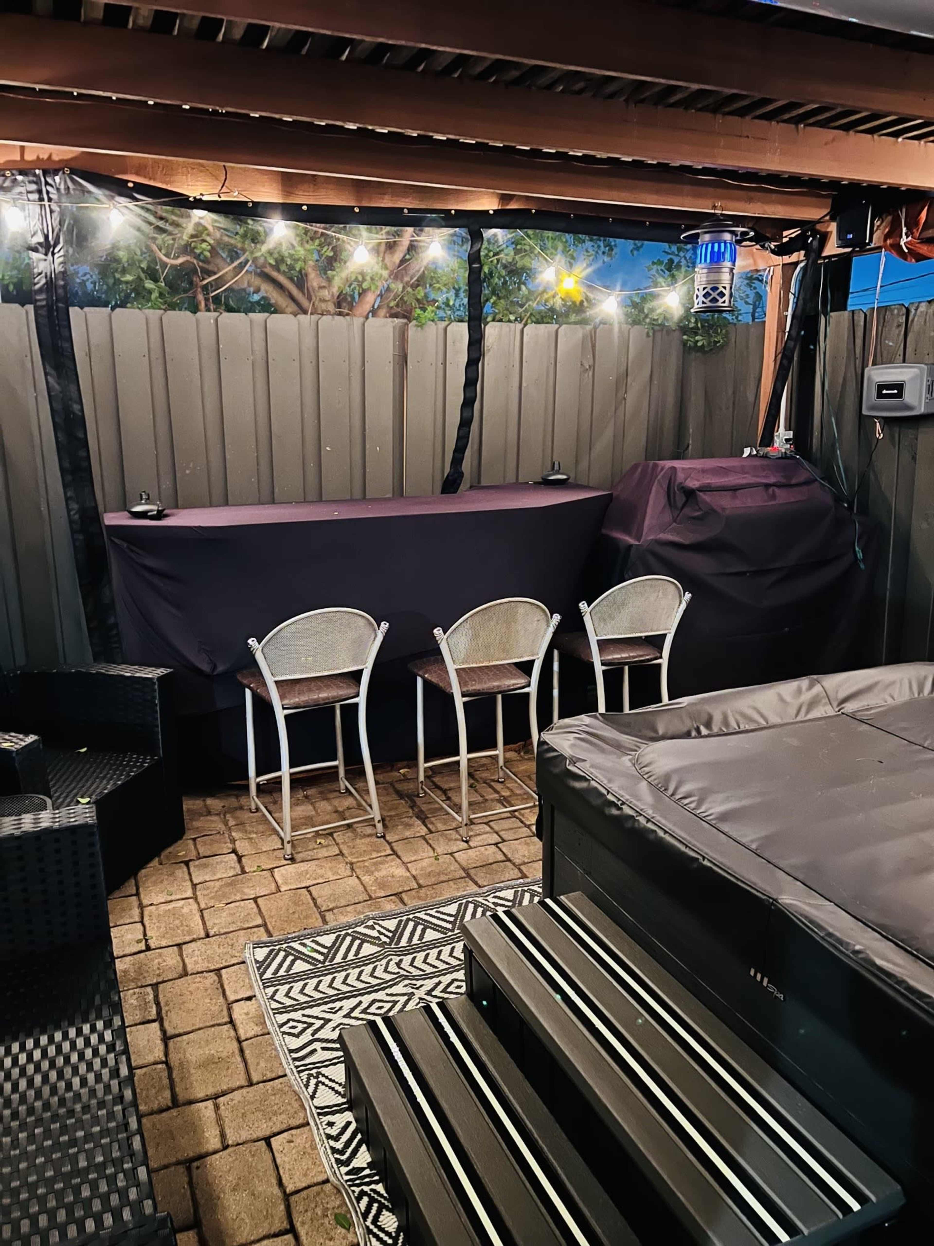 A patio area features a hot tub, a bar counter with three chairs, and a patterned rug on a stone surface, surrounded by wooden fencing and softly illuminated by lights.