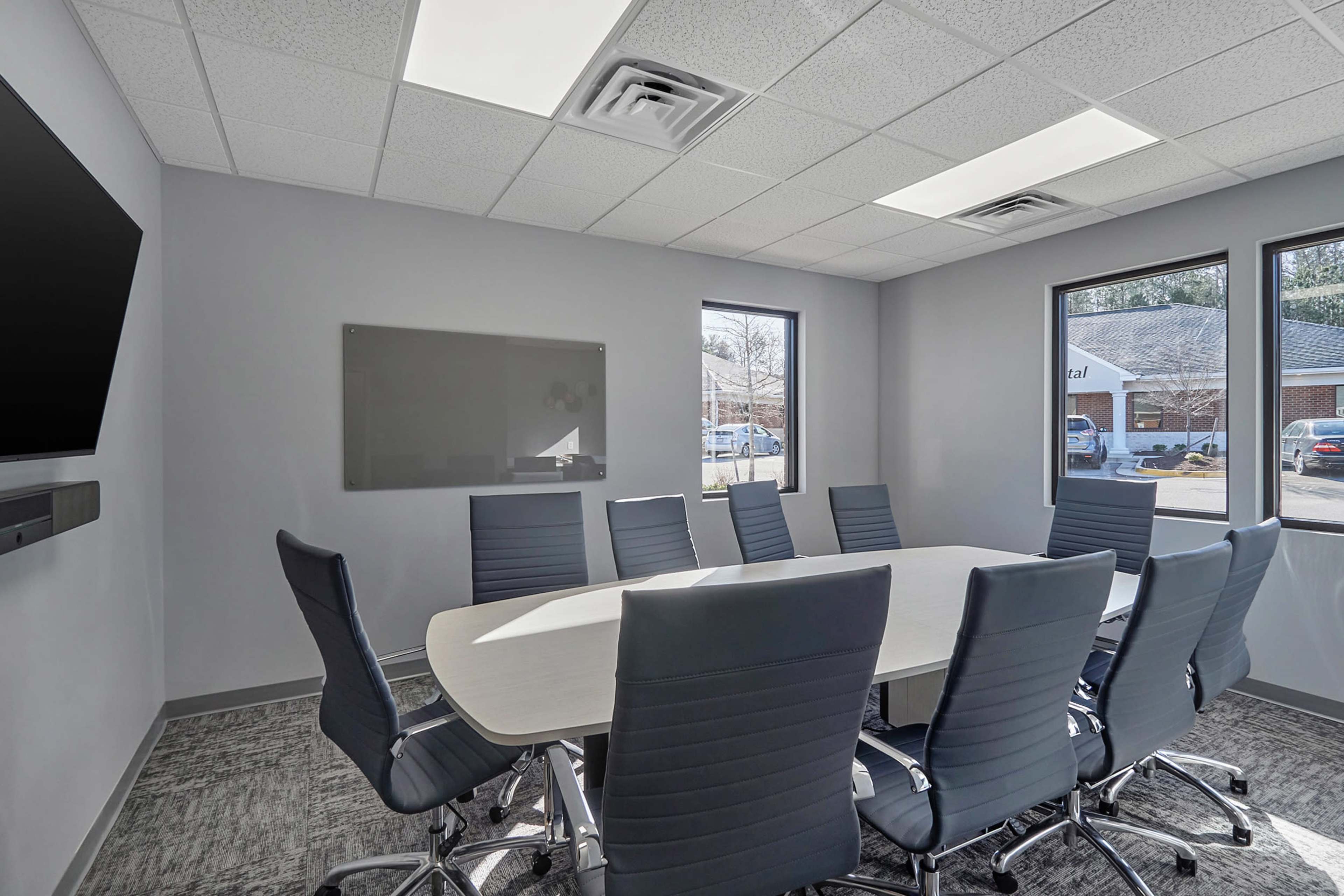 A modern conference room features a large oval table surrounded by ten ergonomic chairs, with windows allowing natural light and a wall-mounted screen.