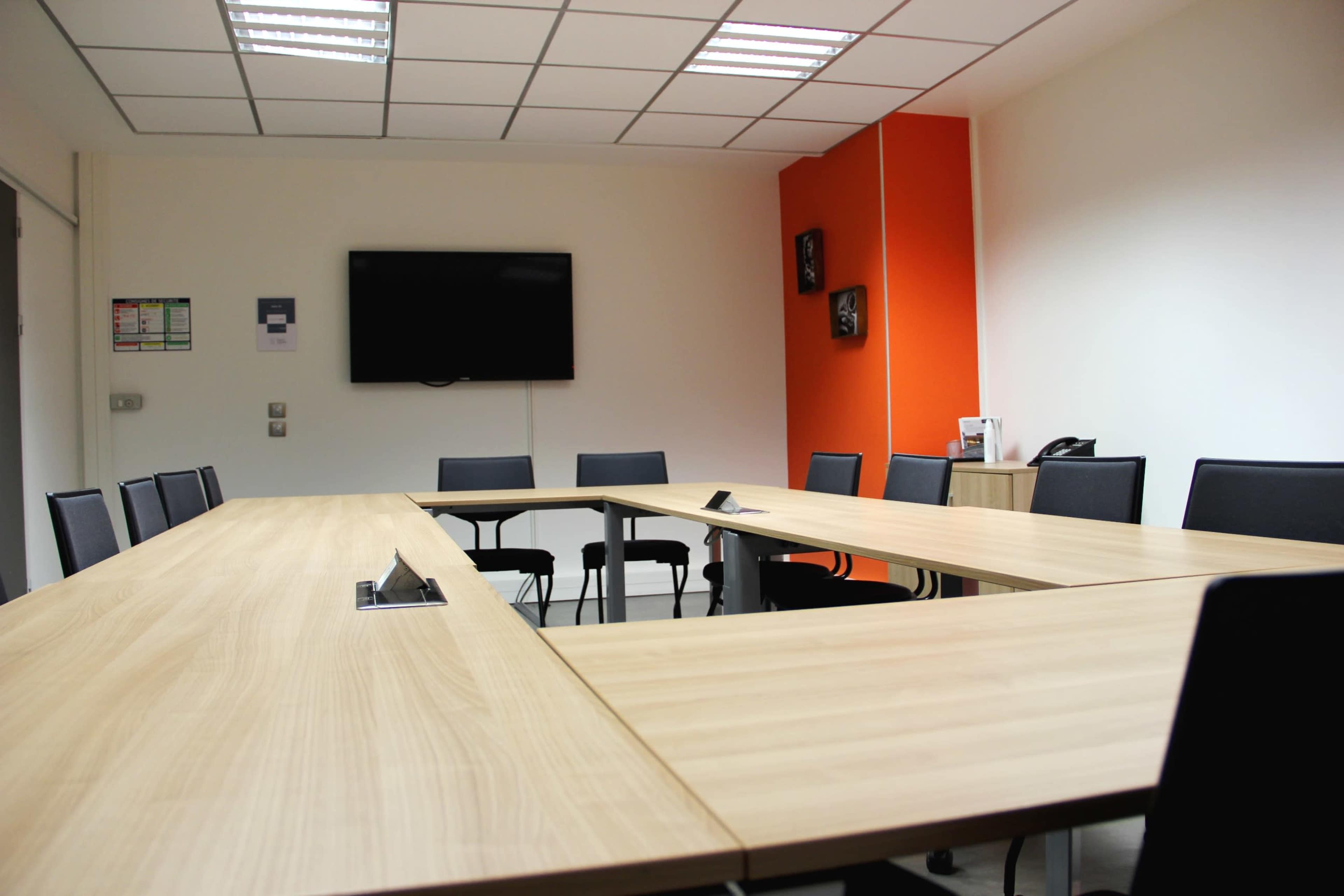A modern conference room arranged with a large wooden table and black chairs, featuring an orange accent wall and a television mounted on one side.