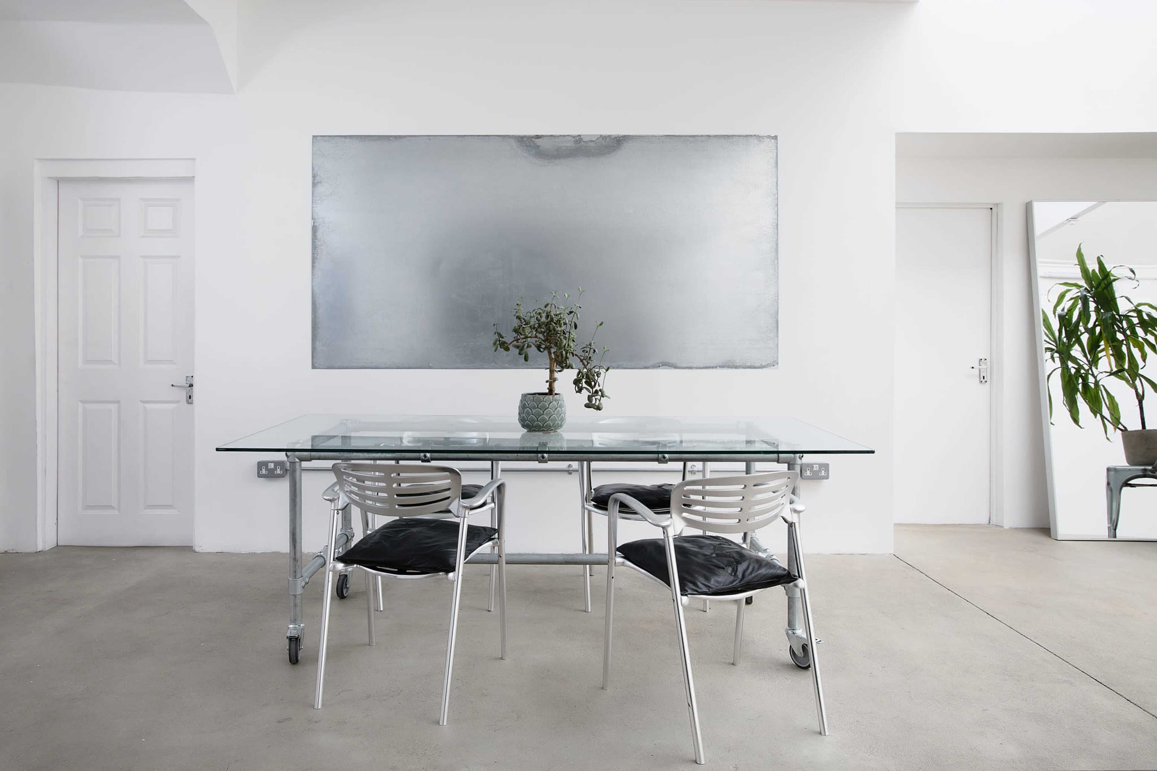 The scene features a minimalist dining area with a glass table, two metal chairs, a potted plant, and a large metal wall art piece against a white background.