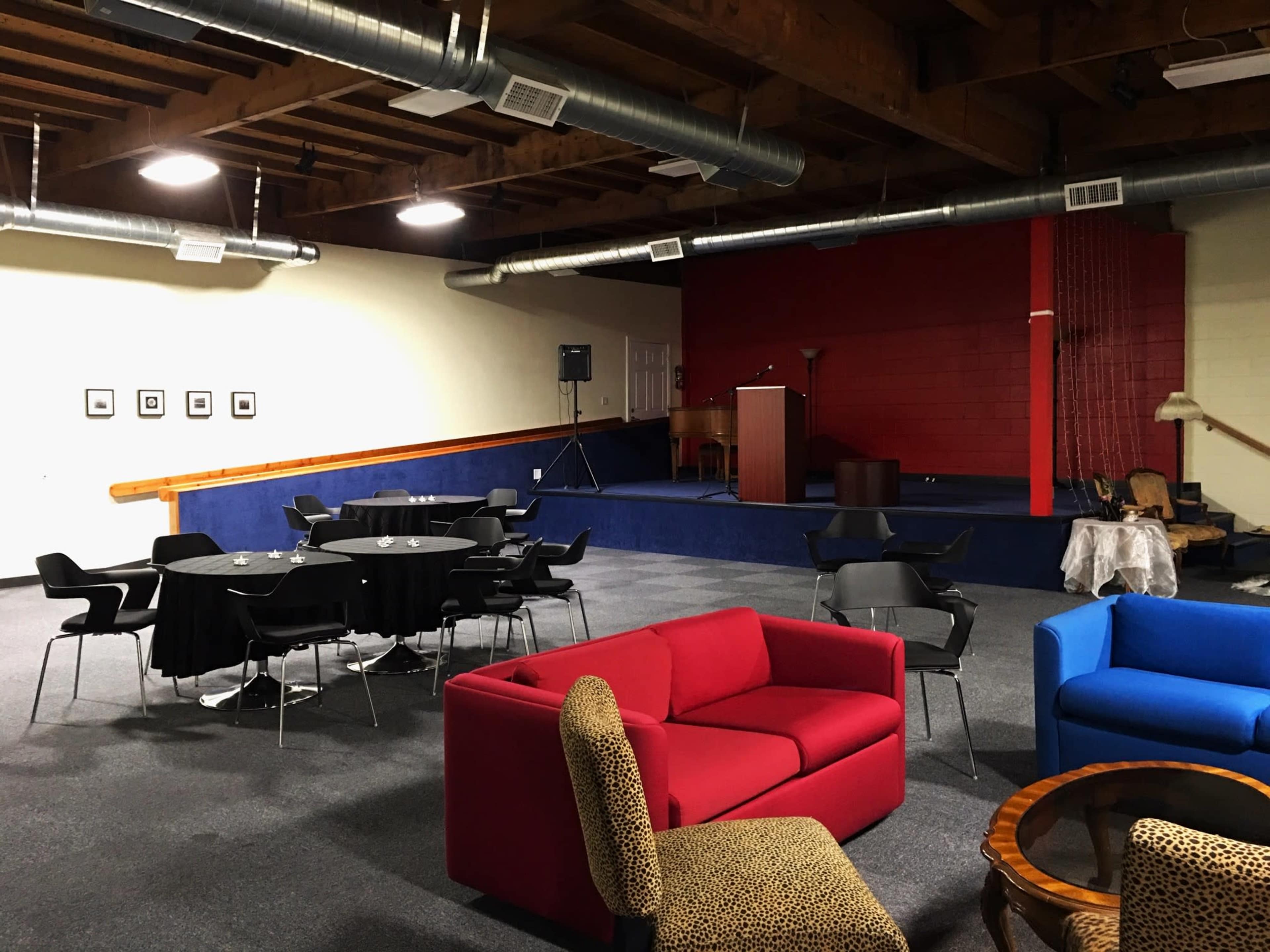 A spacious lounge area with seating arrangements, including black chairs, a red couch, a blue couch, and round tables, adjacent to a small stage.