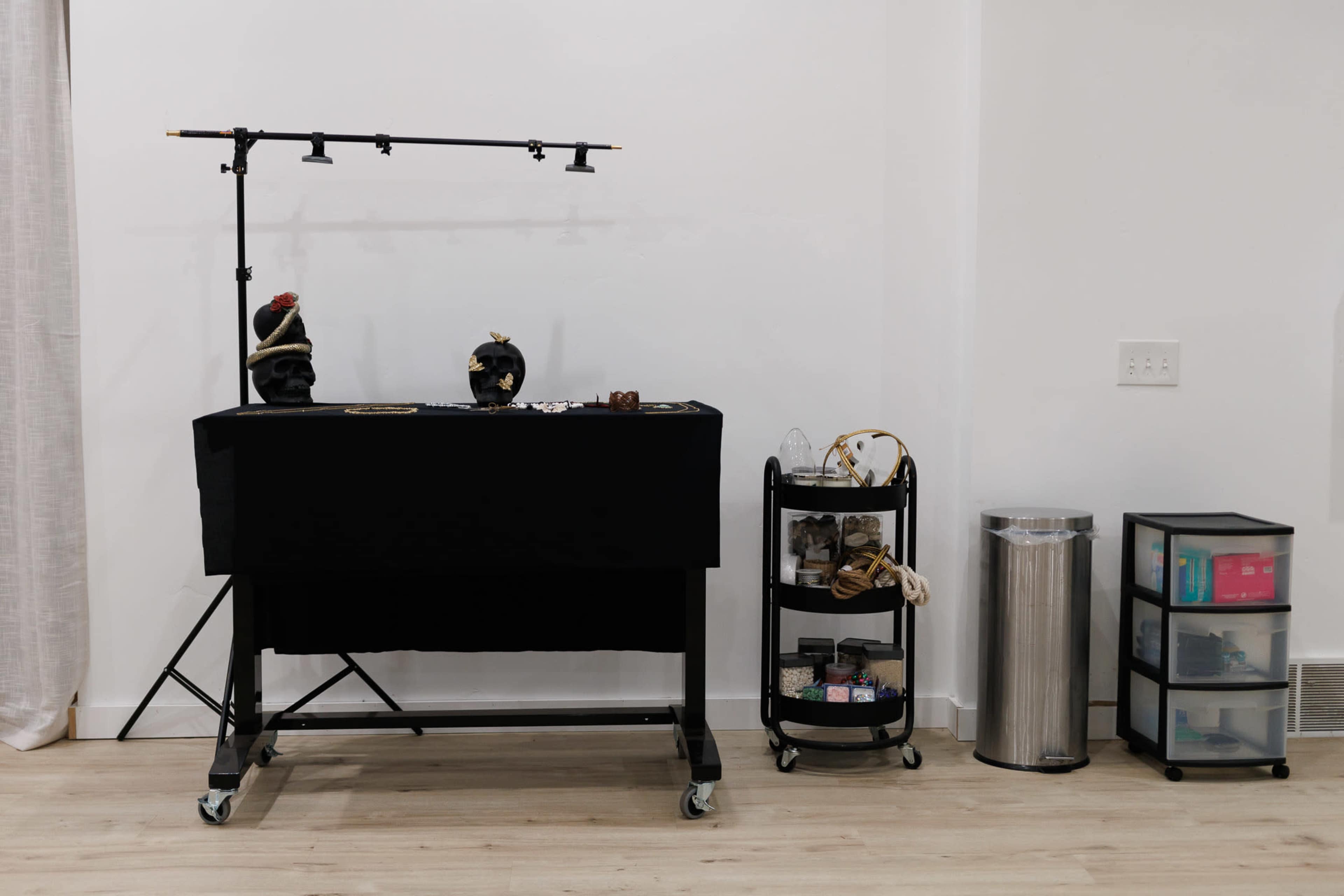 The image shows a display table with black fabric and various items, accompanied by a rolling cart with shoes, a trash bin, and a storage unit against a white wall.